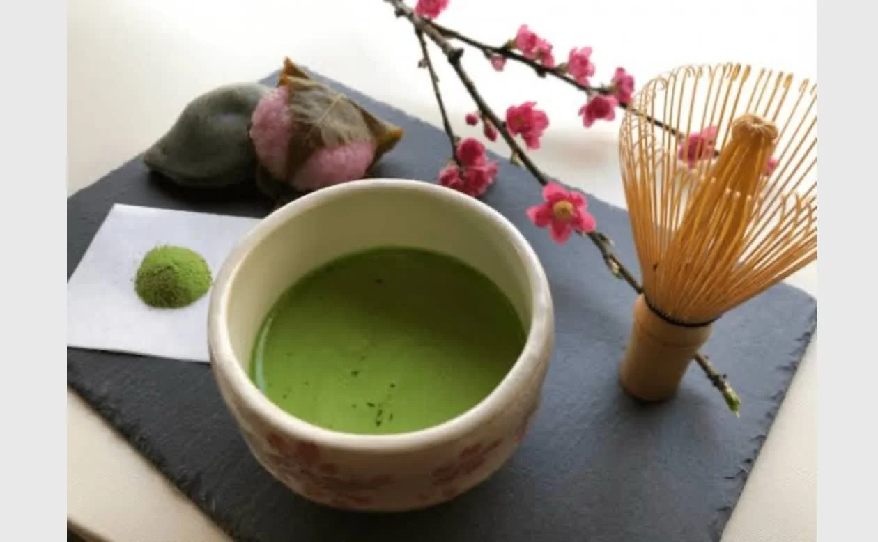 A cup of green matcha tea, a pink and gray decorative cat-shaped object, a small green sphere, pink cherry blossom branch, and a bamboo tea whisk on a slate serving board.