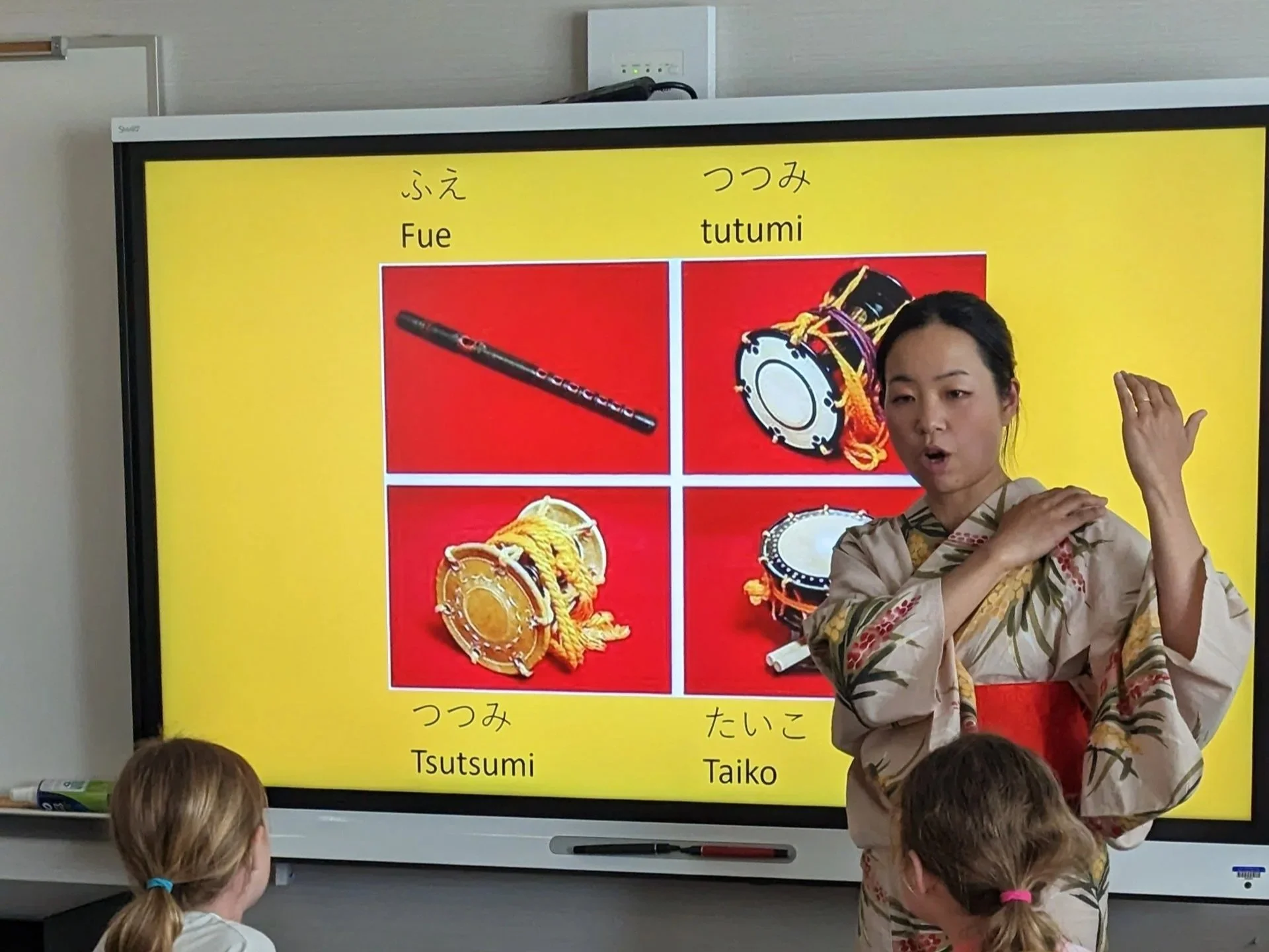 A woman in traditional clothing teaches children about Japanese words for percussion instruments, with images of a flute, drums, and maracas on a bright yellow background