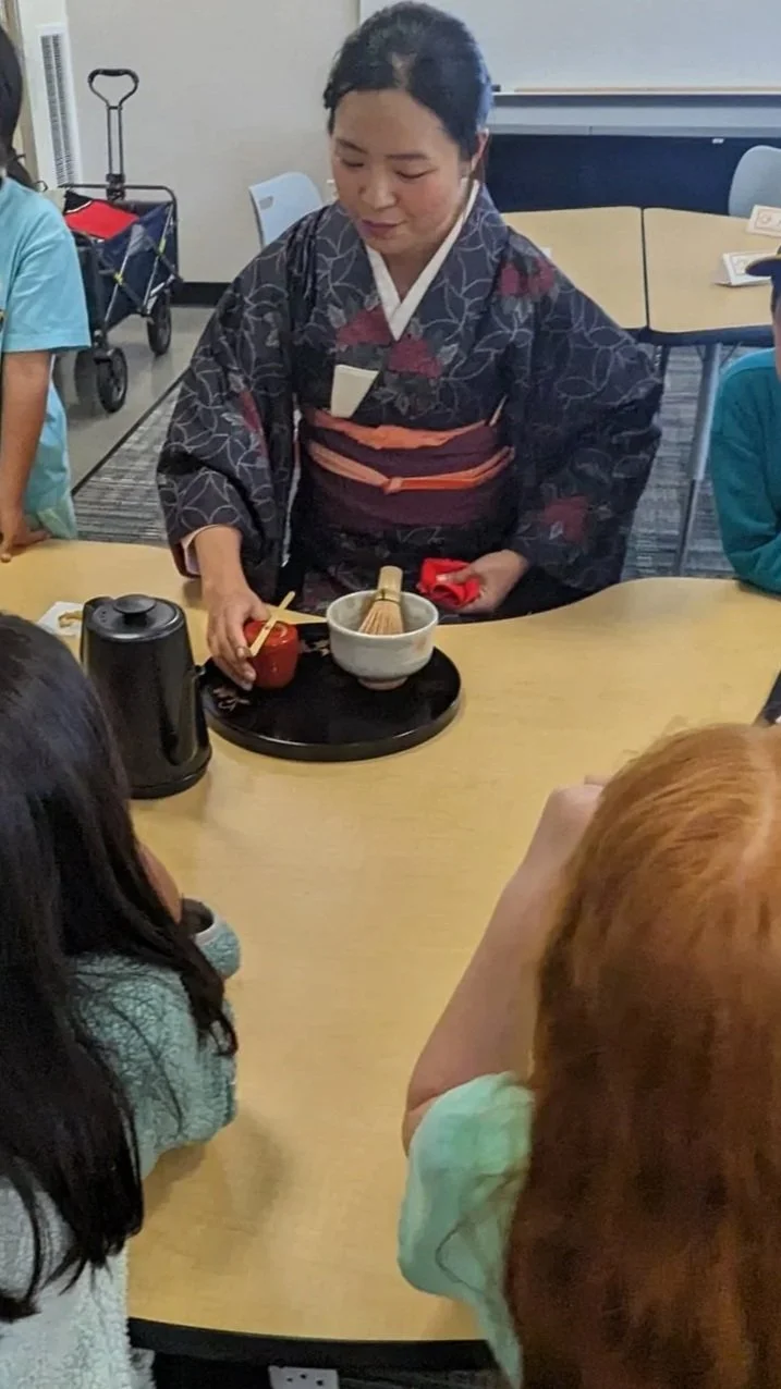 Woman dressed in traditional Japanese kimono performing a tea ceremony for children seated around a table.