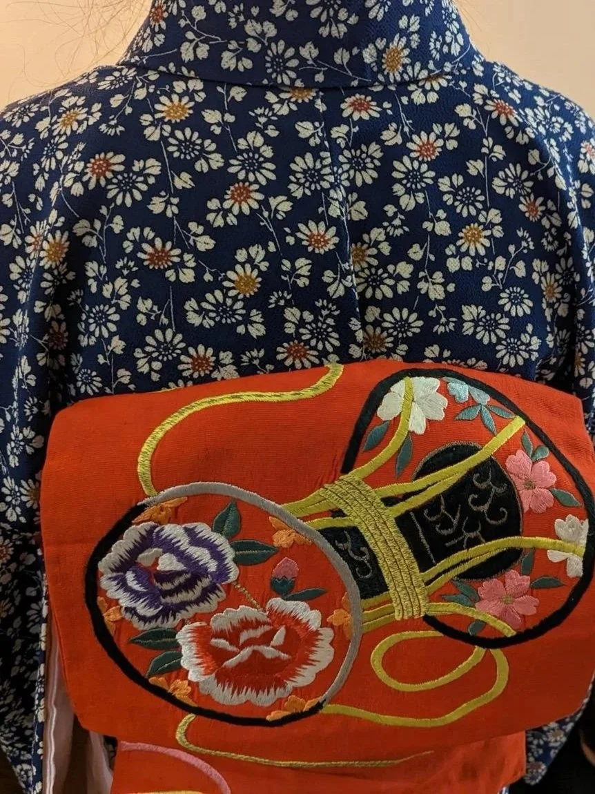 Close-up of the back of a person wearing a traditional Japanese kimono with a navy-blue fabric featuring white and orange floral patterns and an orange obi with intricate embroidery of flowers and a yellow cord.