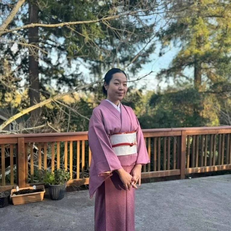 A woman standing outdoors on a wooden deck, wearing a pink traditional Japanese kimono with an obi belt, surrounded by trees and plants.