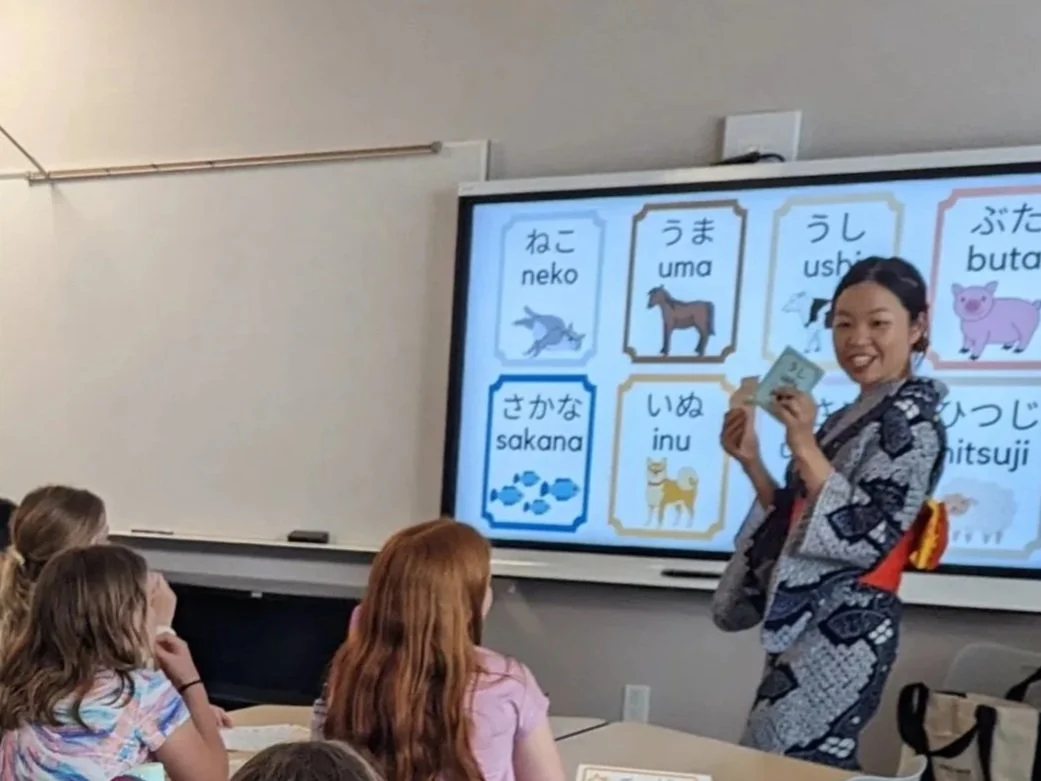 A teacher giving a Japanese language lesson to children in a classroom. The teacher is wearing a traditional kimono, standing in front of a digital display showing Japanese words and pictures of animals. Students are seated at desks, attentively watc