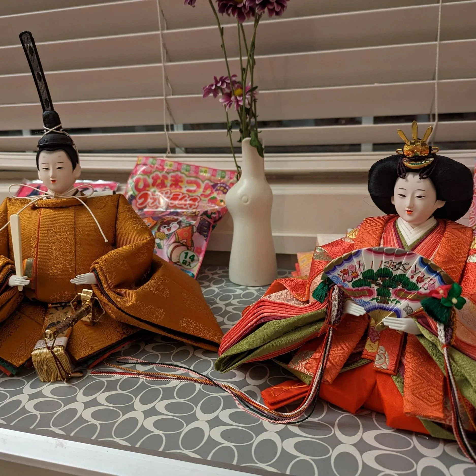 Two traditional Japanese dolls, a male and a female, dressed in elaborate kimono clothing, are displayed on a table with a patterned surface, in front of window blinds. A white vase with pink and purple flowers and some Japanese snack packaging are a
