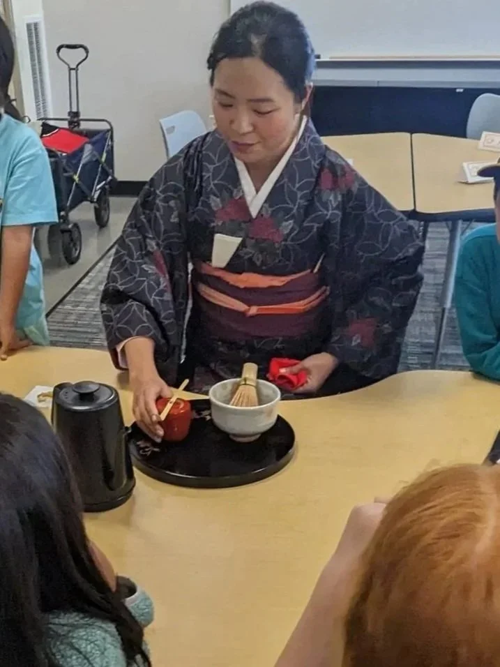 Woman dressed in traditional Japanese kimono performing a tea ceremony for children seated around a table.