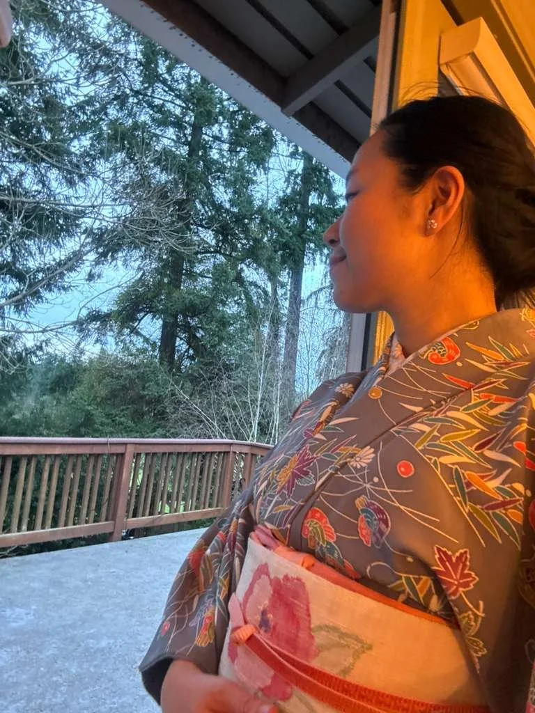 A woman wearing a traditional kimono stands on a balcony, looking outside at trees during dusk.