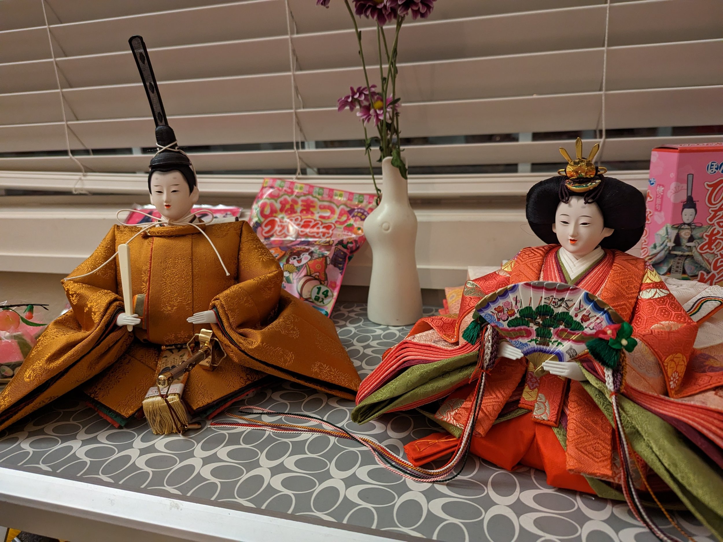 Two traditional Japanese dolls, a male and a female, dressed in elaborate kimono clothing, are displayed on a table with a patterned surface, in front of window blinds. A white vase with pink and purple flowers and some Japanese snack packaging are a