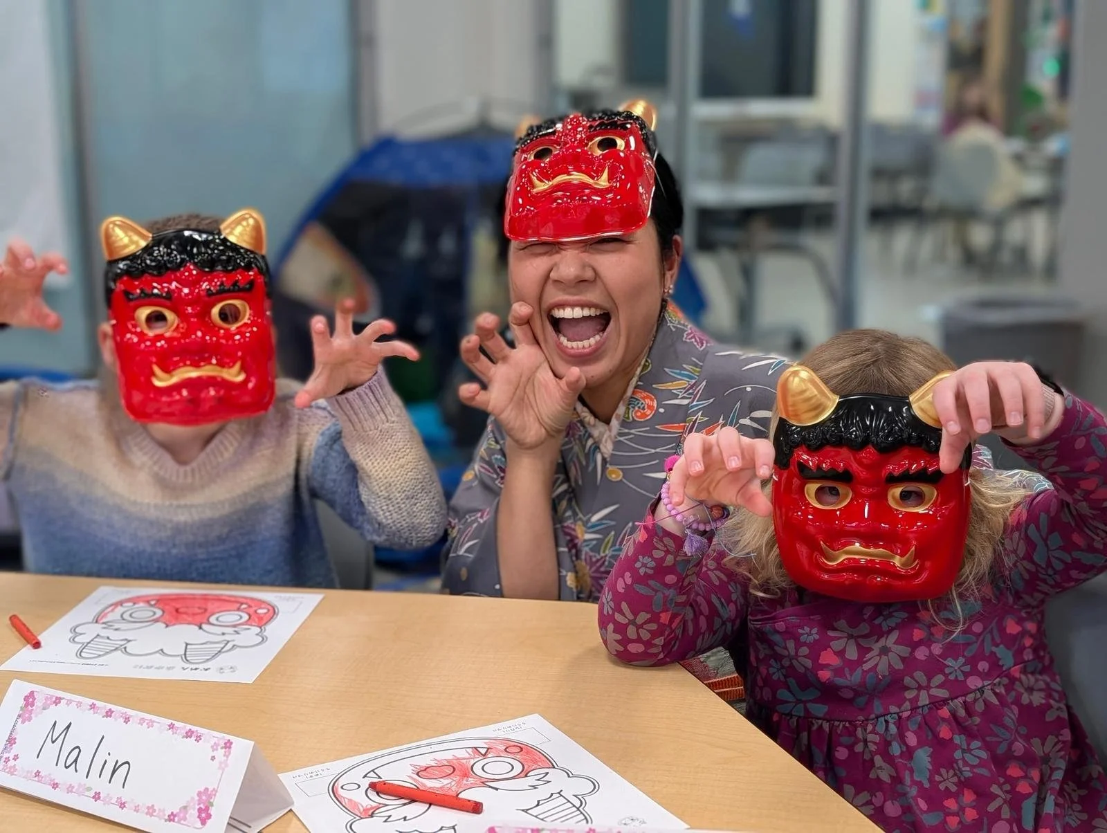 Three people, two children and one adult, wearing red and black traditional demon masks and making tiger claw gestures at a table with coloring pages. The children are seated and the adult is standing, all smiling and playing.