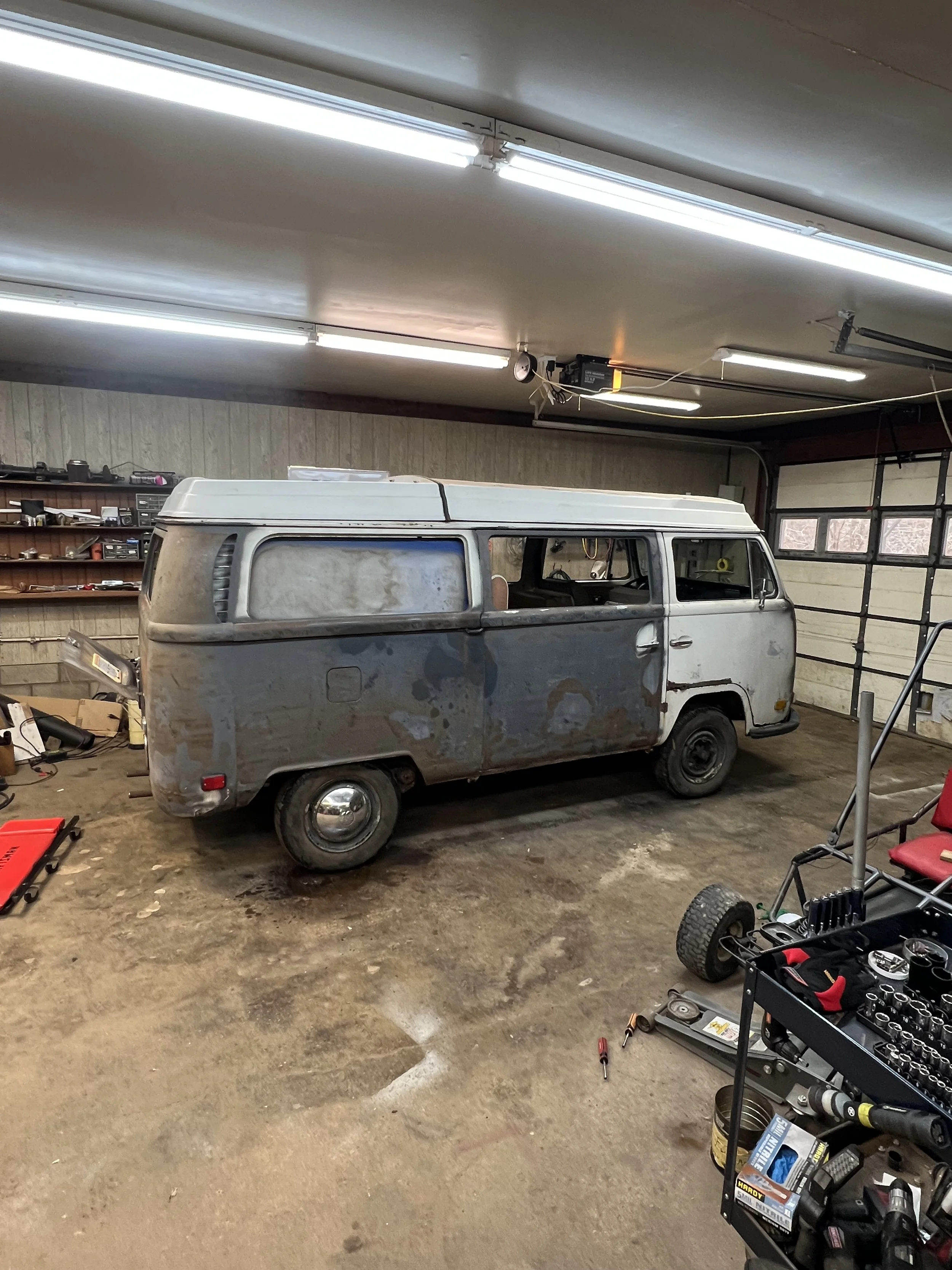 A rusted, vintage Volkswagen Type 2 van in a garage with tools and equipment around.