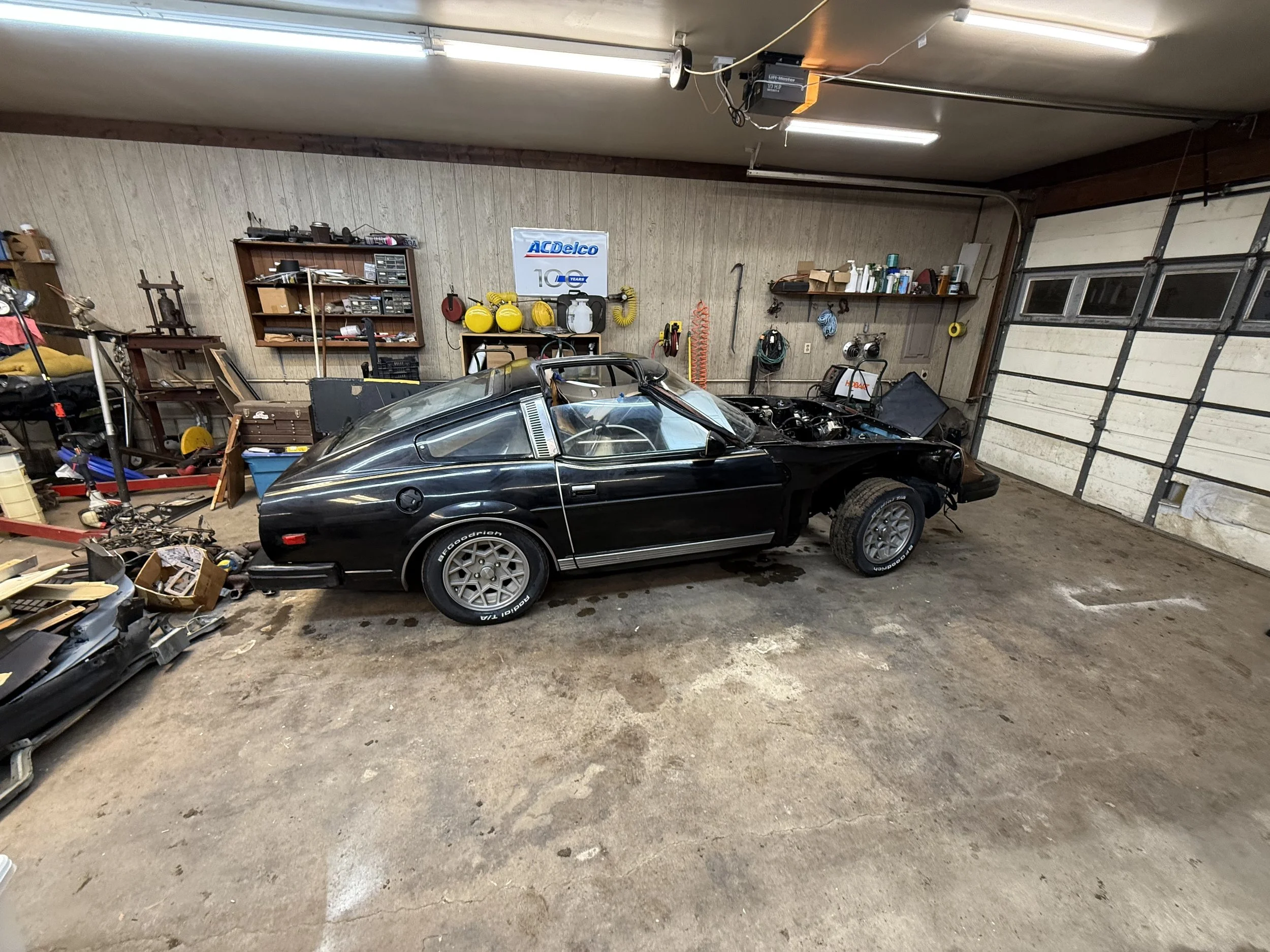 A partially disassembled black sports car in a cluttered garage with various tools, shelves, and equipment.