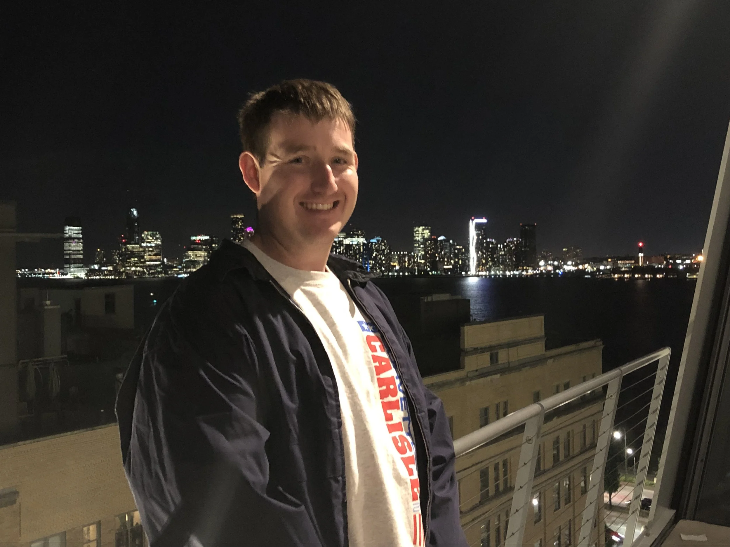 A smiling man standing on a balcony at night with a city skyline illuminated in the background.