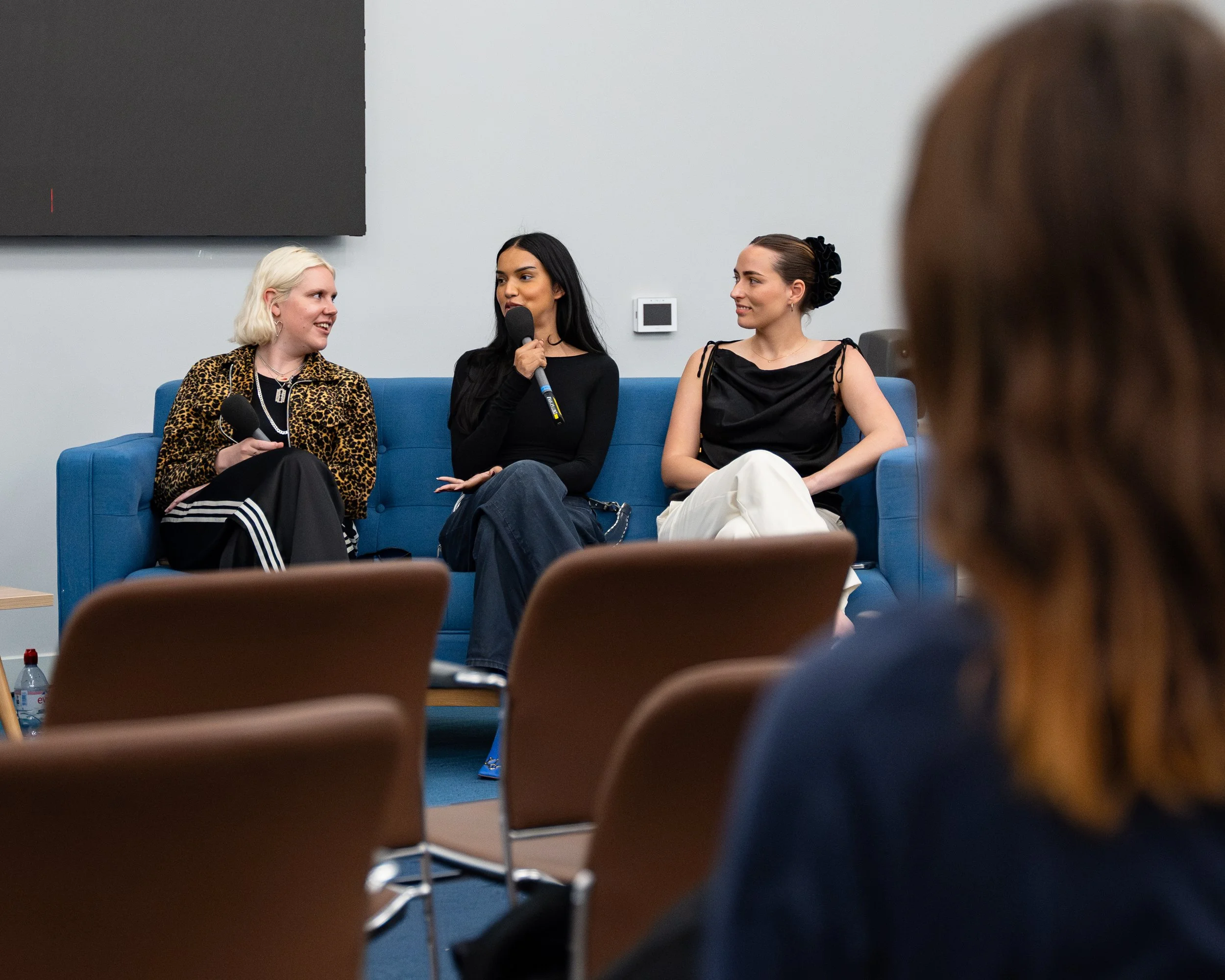 Three women seated on a blue couch participating in a panel discussion, with an audience in the foreground, microphones in hand, and a large screen behind them.