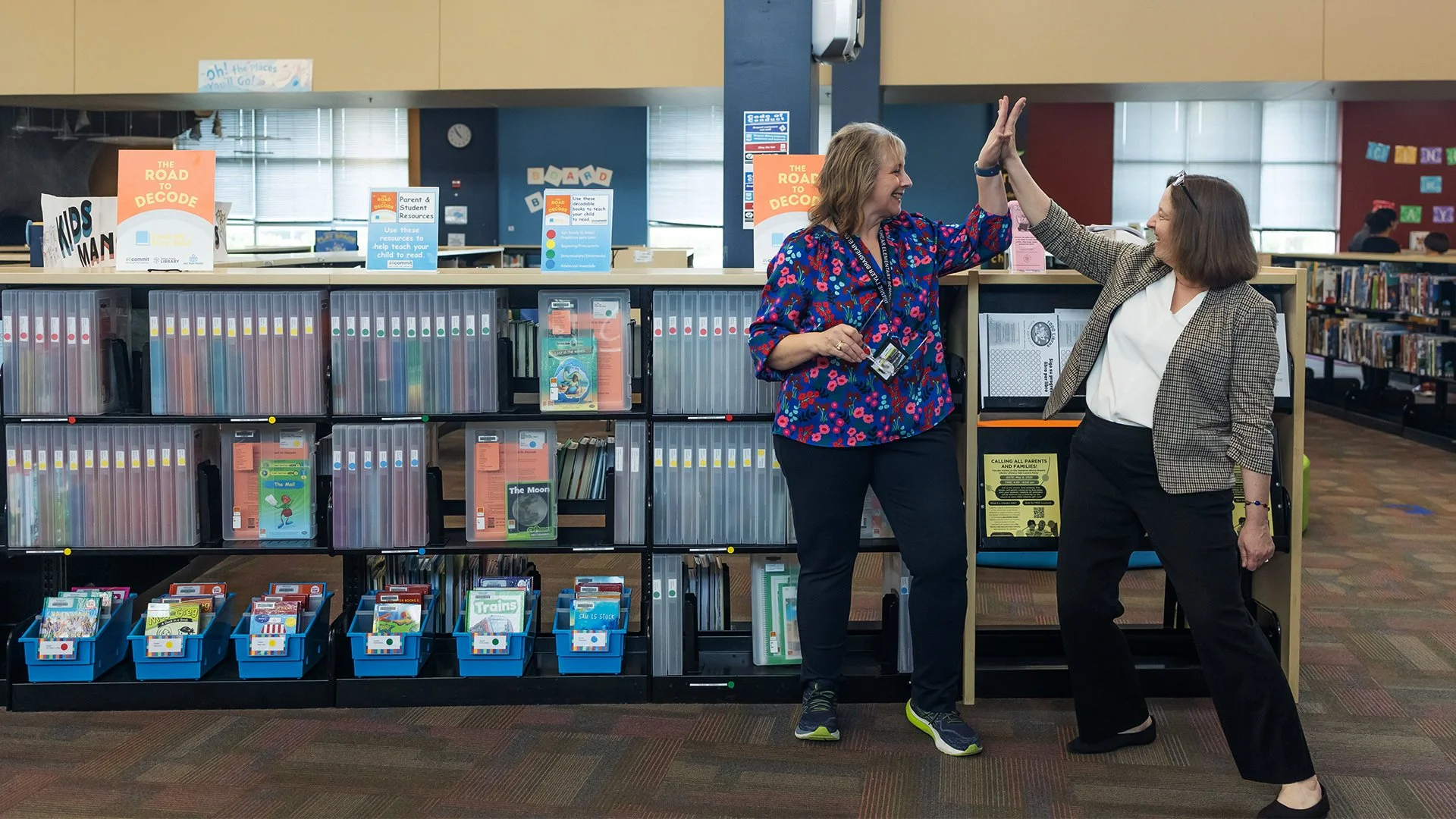Two women are giving each other a high five in a library. They are standing in front of bookshelves filled with books and educational materials.