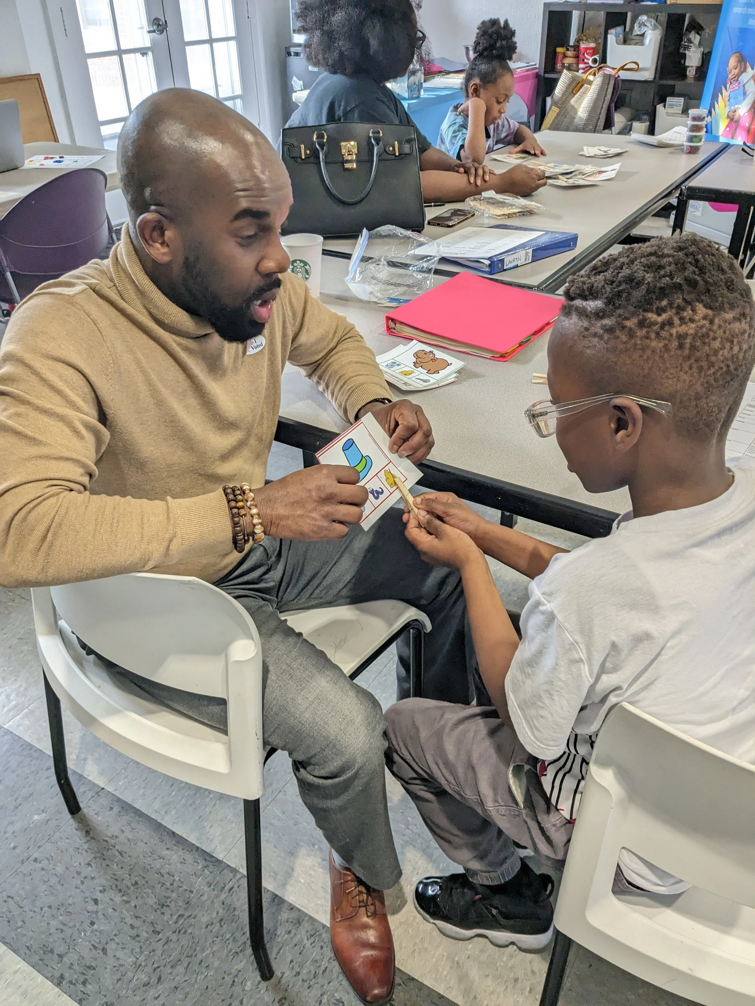 A man and a boy are sitting at a table, engaging in a learning activity involving picture cards. The man is holding a card with a banana, and the boy is pointing at the card with a small stick. There are various papers, folders, and beverages on the table, with other children and adults in the background participating in activities.