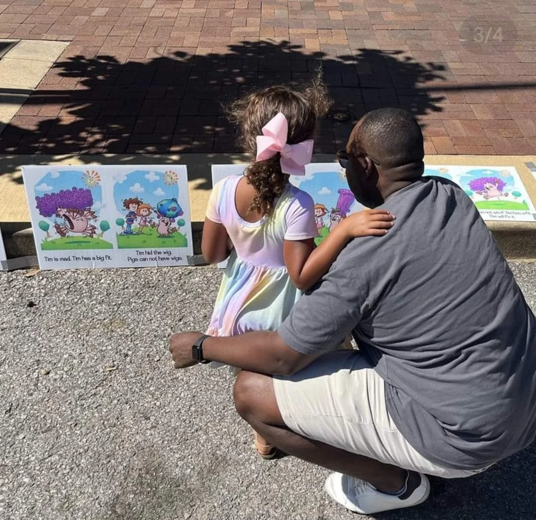 A man and a young girl are looking at colorful illustrated storybook pages displayed outside on a low ledge. The girl has a big pink bow in her hair and is wearing a light-colored dress, while the man is wearing a gray shirt, white shorts, and a black face mask. It is sunny and the scene is shaded by nearby trees.