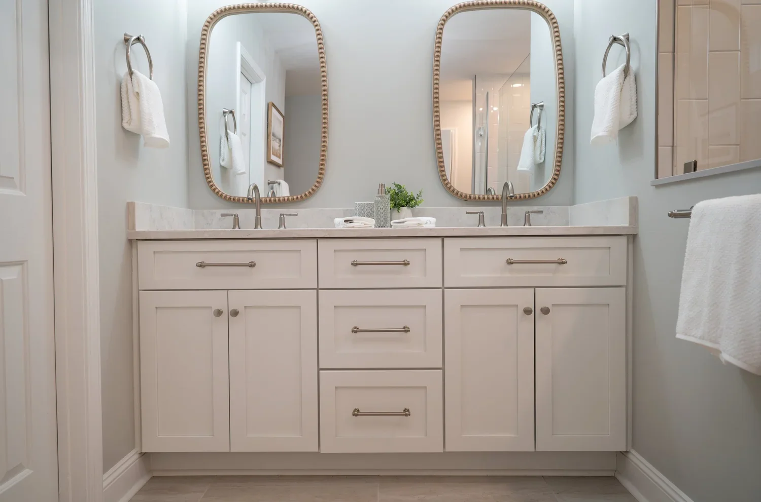 Bathroom vanity with two mirrors, a white cabinet, and two sinks, decorated with small plant and towels.