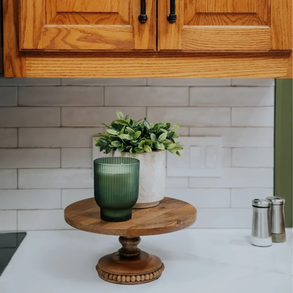 A kitchen countertop with a small wooden cake stand holding a potted green plant and a green glass candle holder, with a white brick backsplash and wooden cabinets above.