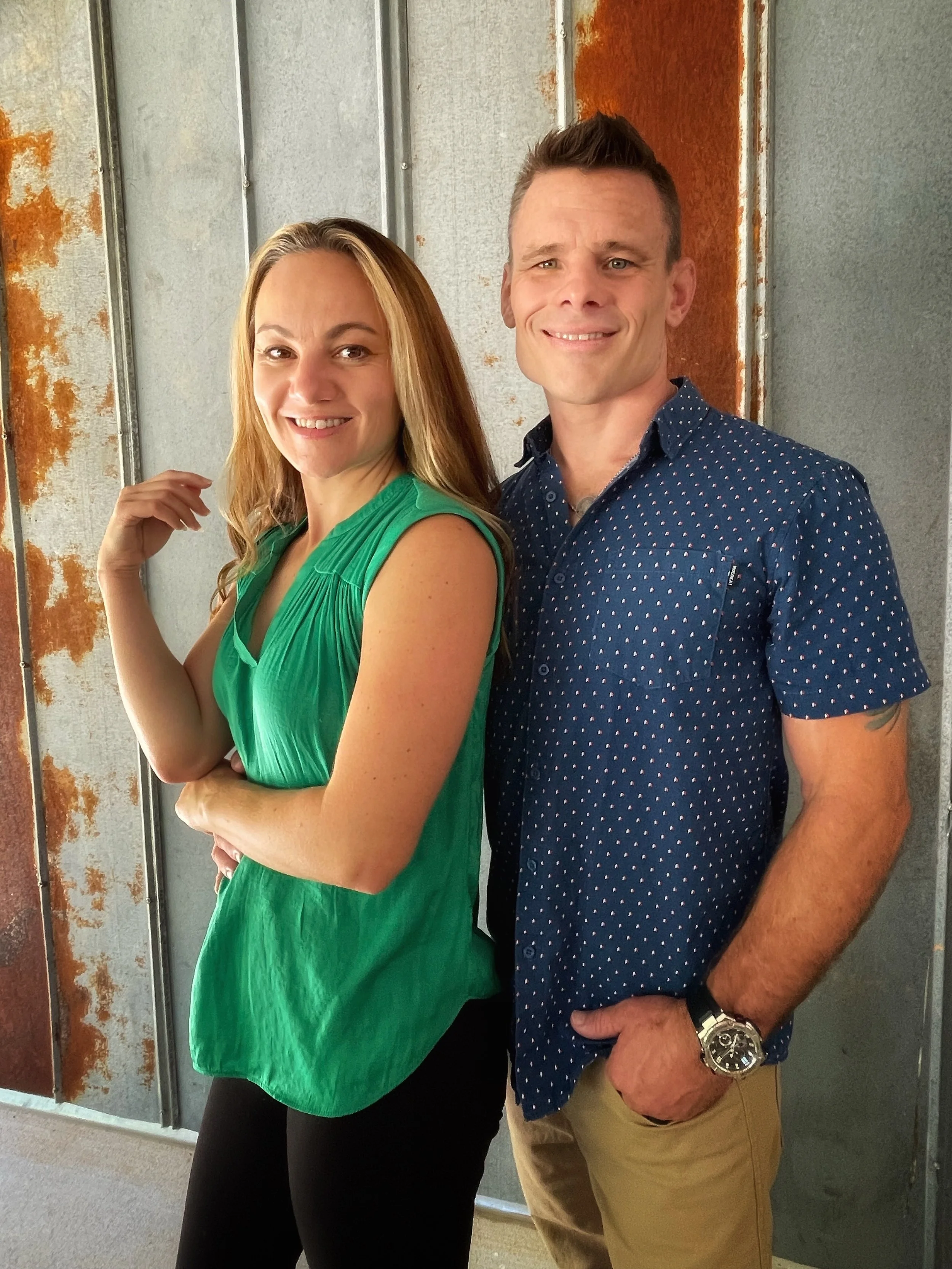 A smiling woman in a green sleeveless top and a man in a blue patterned shirt standing in front of a rusted metal wall.