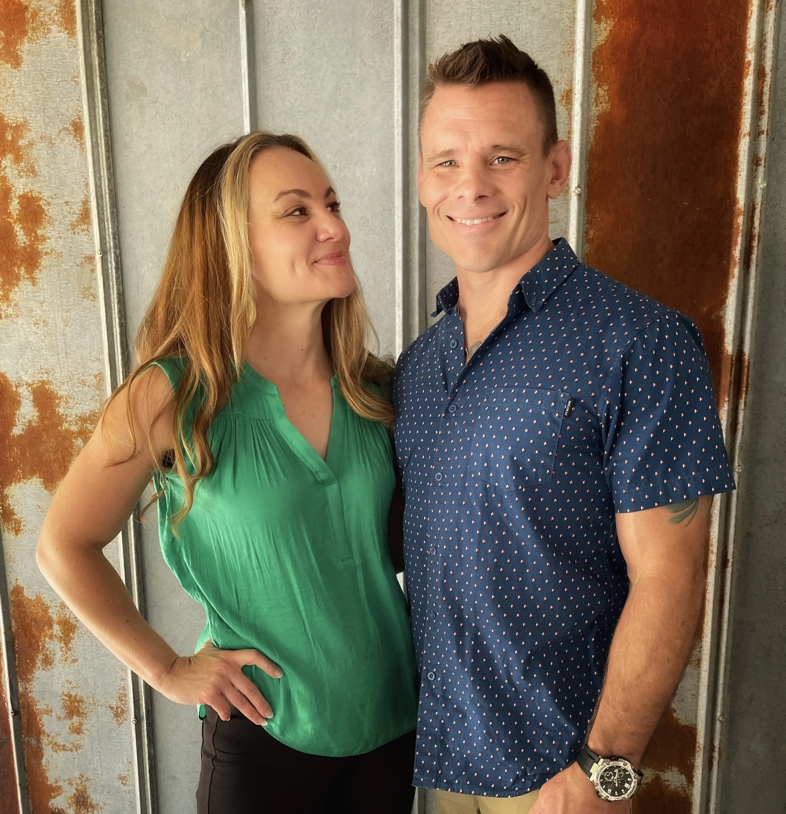 A woman with long red hair and a man with short brown hair smiling at each other, standing against a rustic metal wall.