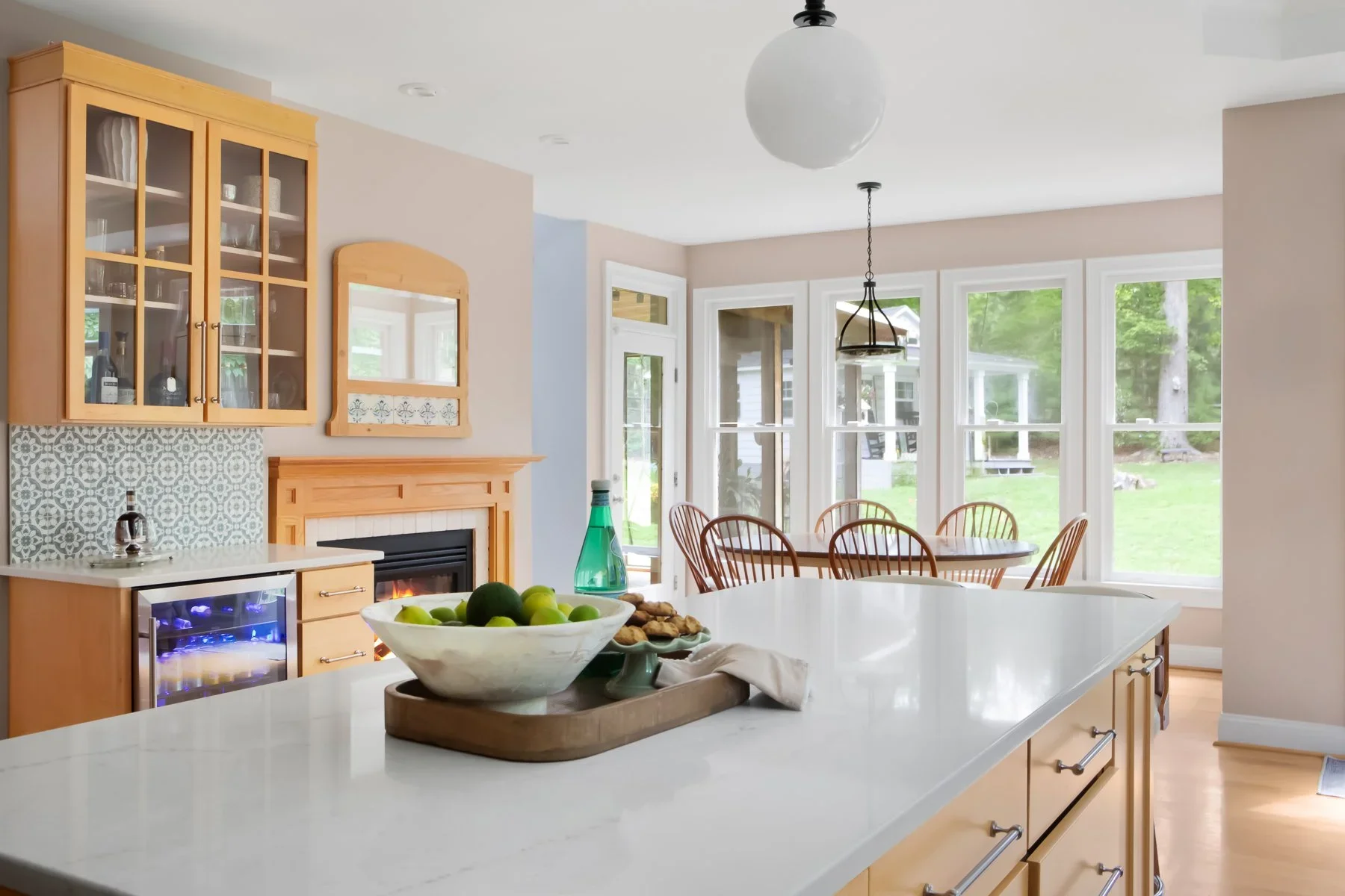 Bright, airy kitchen with large island, bowl of green apples, and adjacent dining area with wooden table and chairs, large windows revealing green outdoor space.