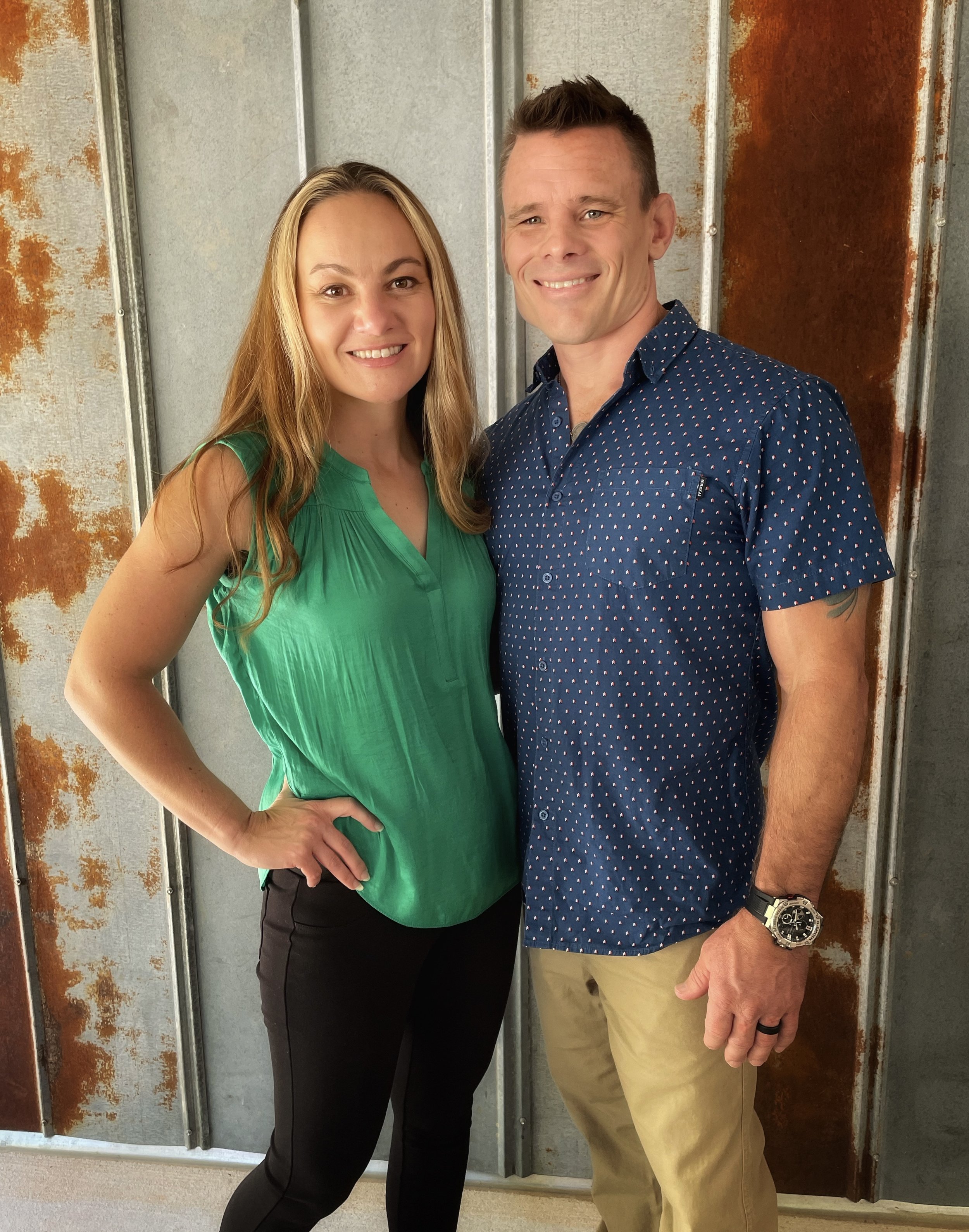 A man and woman stand closely together, smiling at the camera, against a rustic, metal wall background.