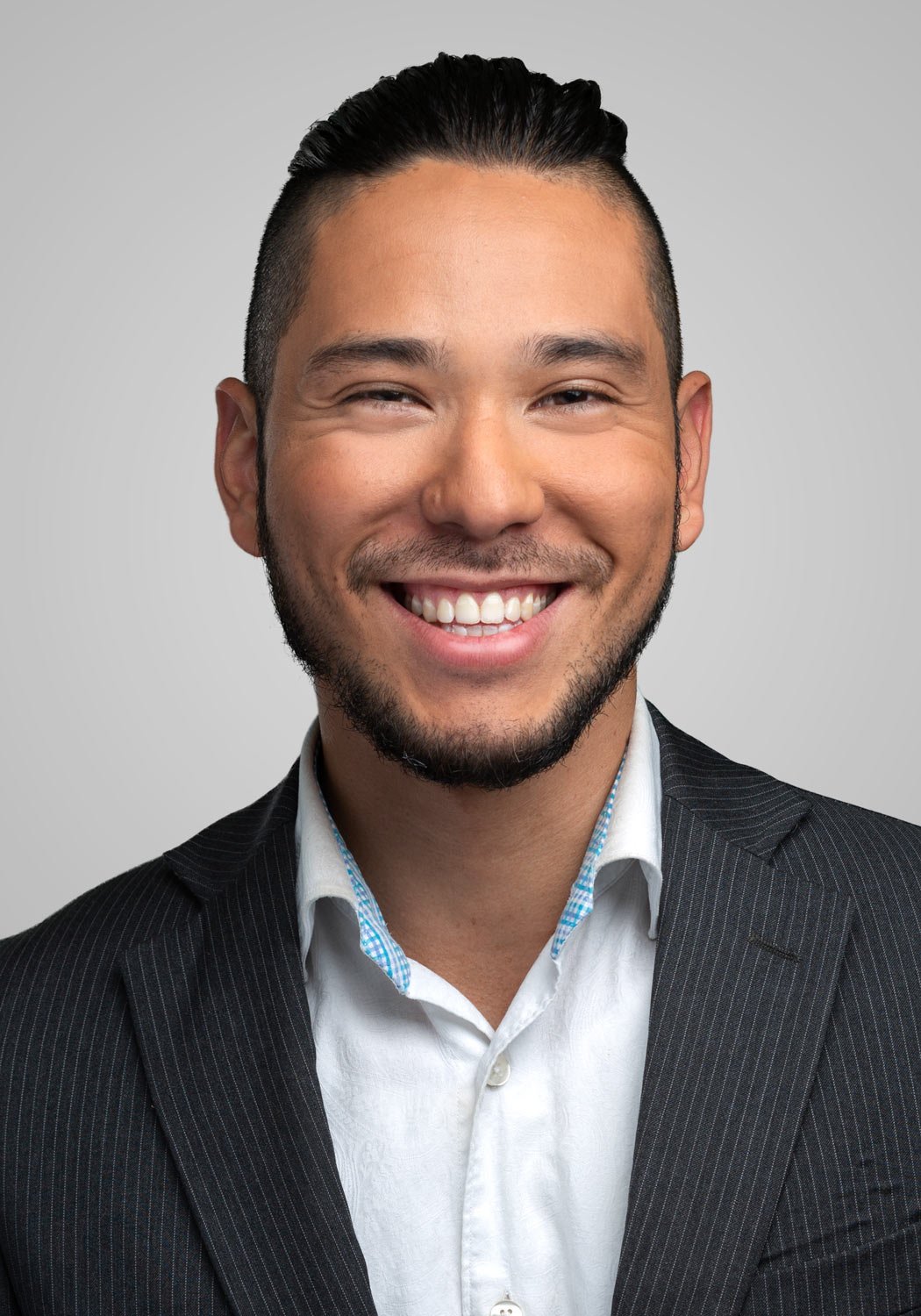 Portrait d'un homme souriant avec une barbe, portant un costume foncé et une chemise blanche à col boutonné, fond gris clair.