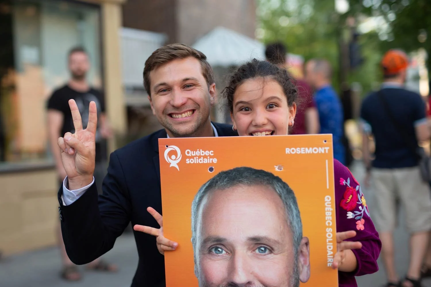 Un homme et une jeune fille posent avec un panneau de campagne politique lors d'un rassemblement en plein air. L'homme fait un signe de paix avec la main. La scène semble être en Québec, vulnérable à un climat de campagne.