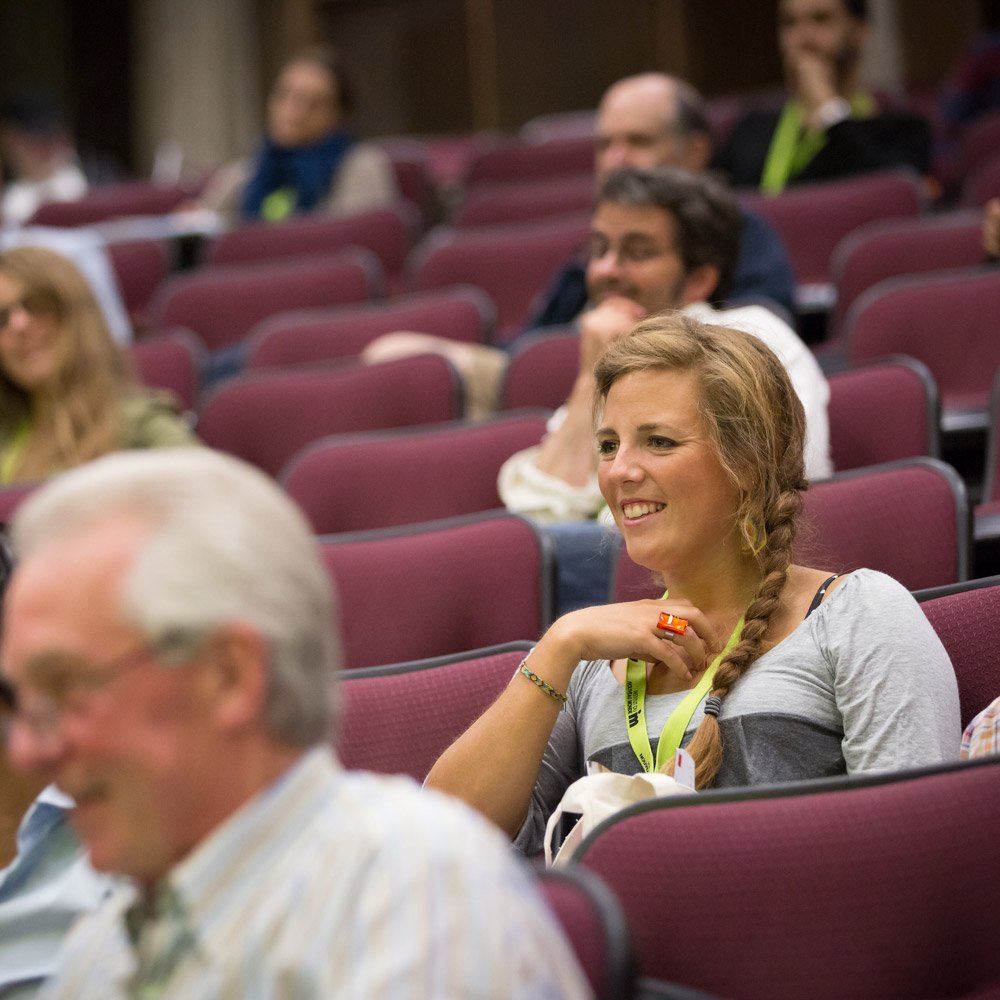 Une femme souriante assise dans une salle de conférence ou théâtre, entourée de plusieurs autres personnes attentives, toutes assises sur des sièges rouges.