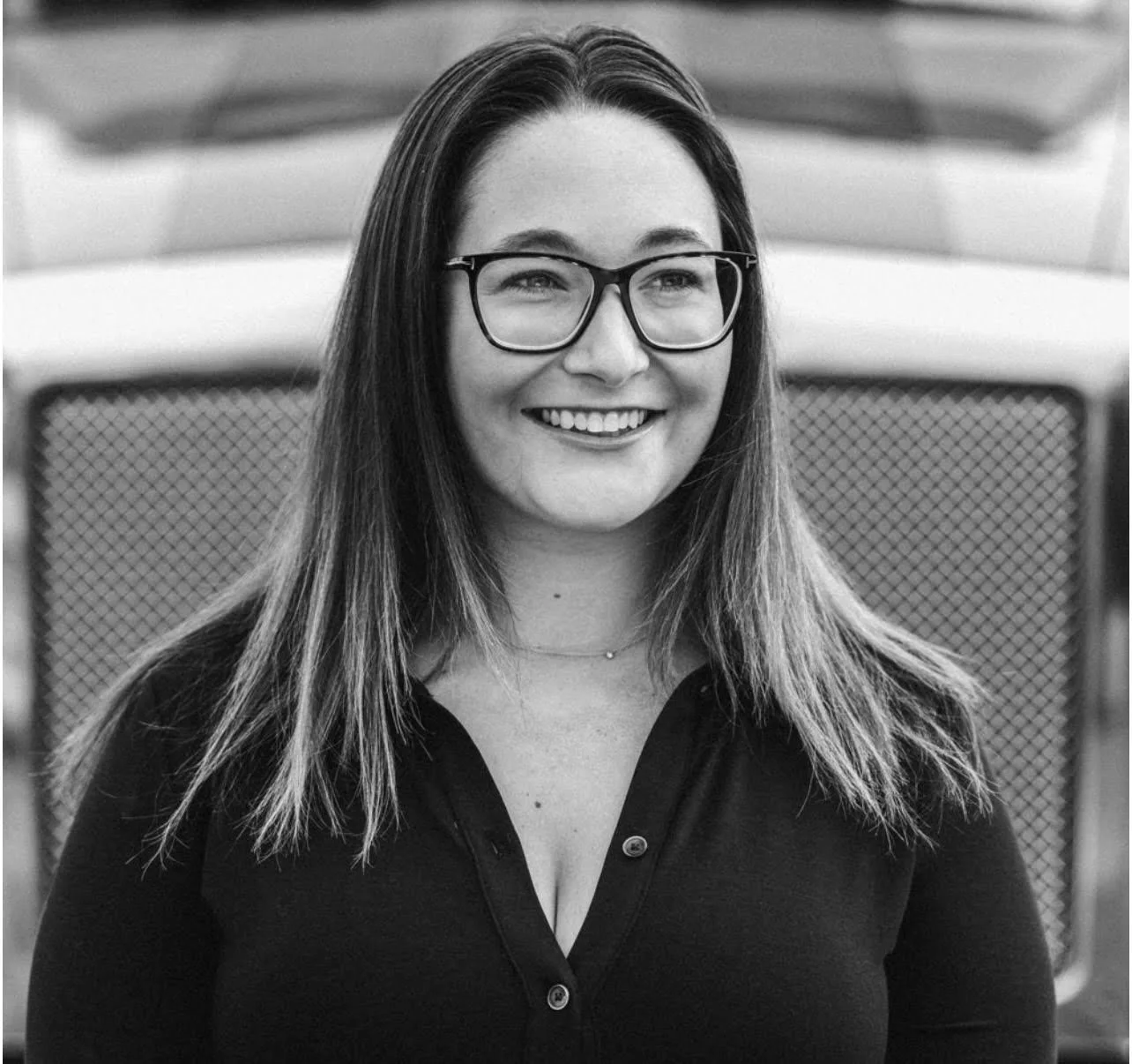 A smiling young woman with glasses and long hair, wearing a dark top, in front of a vintage car.