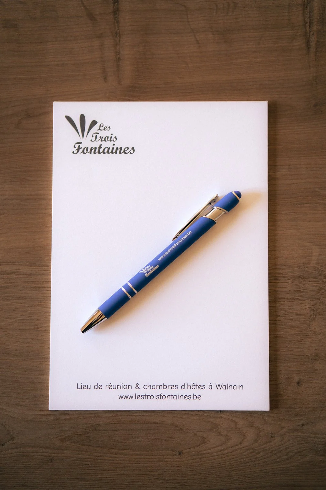 Une feuille blanche avec le logo 'Les Trois Fontaines' en haut à gauche, un stylo bleu posé dessus, et du texte en bas indiquant le lieu de réunion et le site web.