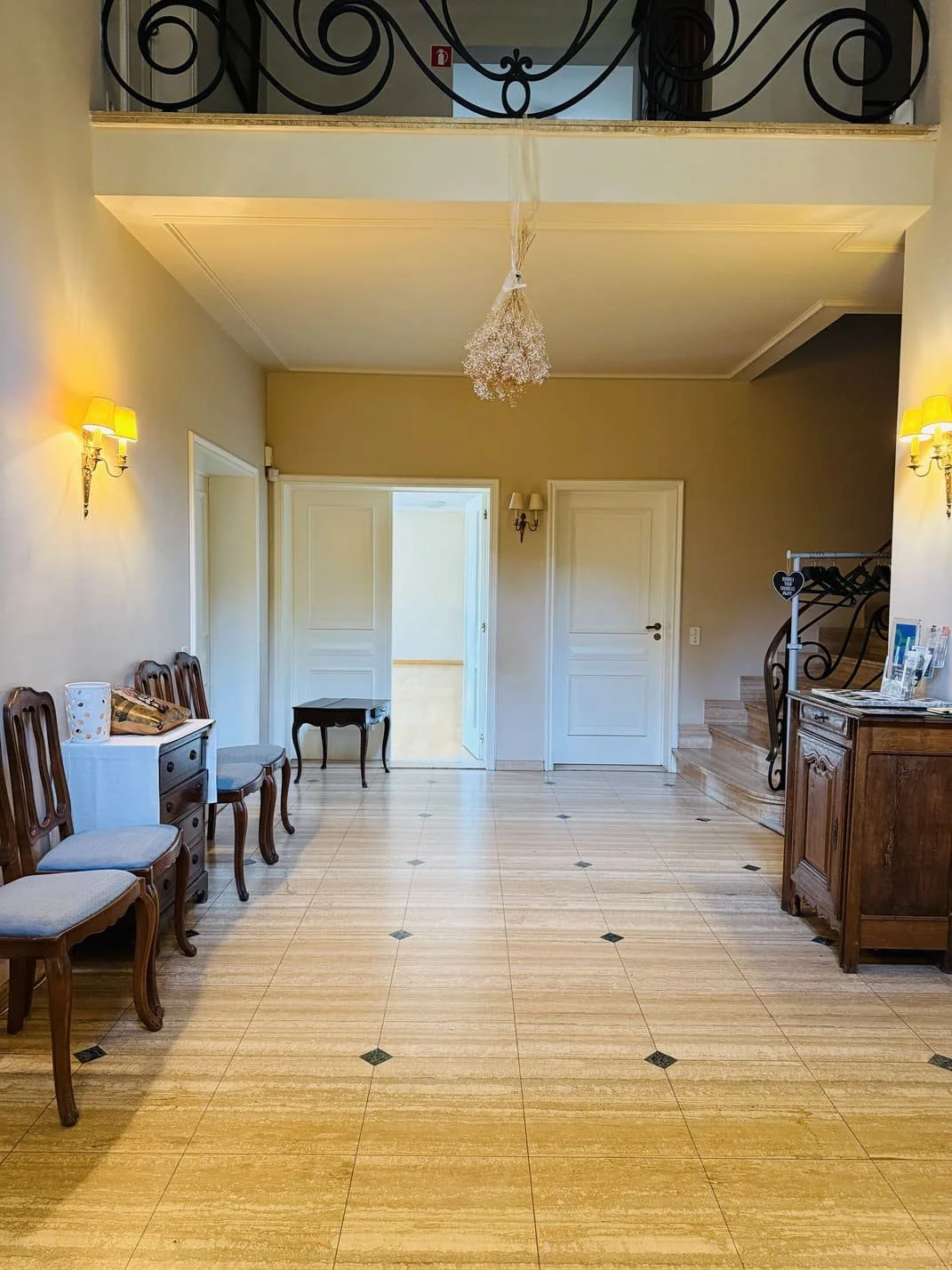 Intérieur d'un hall d'entrée avec des chaises en bois et tissu beige, un meuble en bois, des lampes murales, un escalier en colimaçon, et une porte à l'arrière. Décor classique et chaleureux.