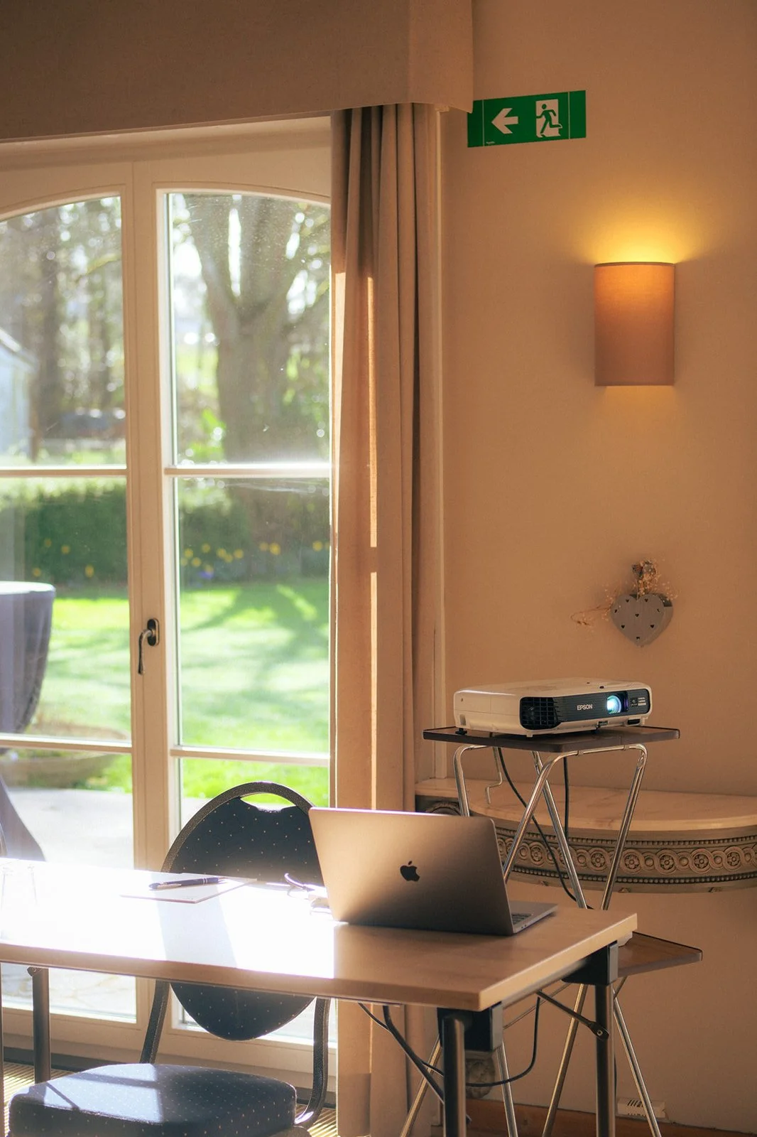 Un espace de réunion lumineux avec une porte fenêtre donnant sur un jardin. Sur la table, un ordinateur portable, à côté un projecteur sur une petite table. Au mur, un éclairage chaud et une décoration en forme de cœur.