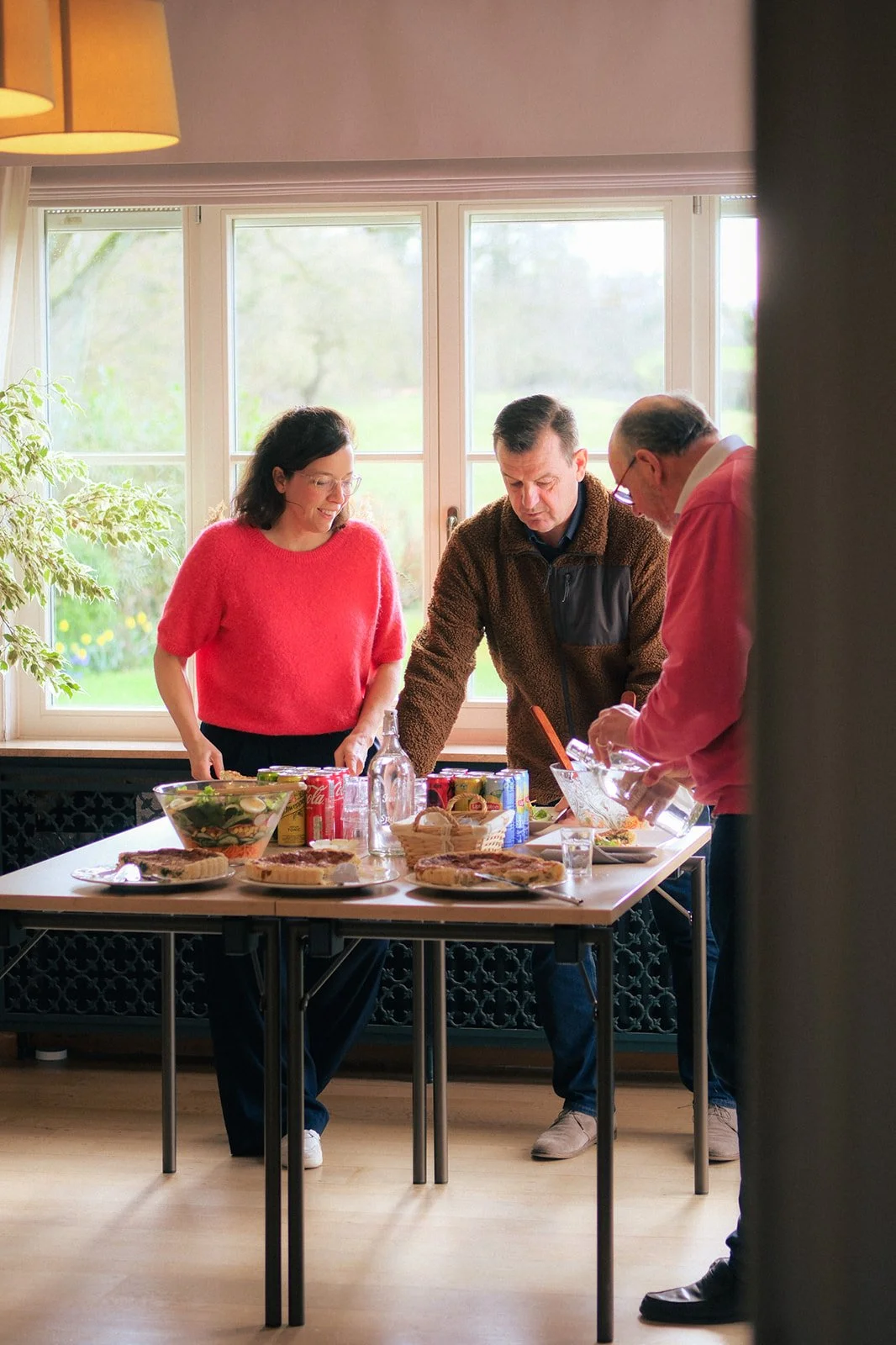 Trois participants se servent au buffet pendant la pause de la réunion dans une pièce lumineuse avec une grande table. Il y a des quiches, des salades, des sodas, et des bouteilles d'eau sur la table.