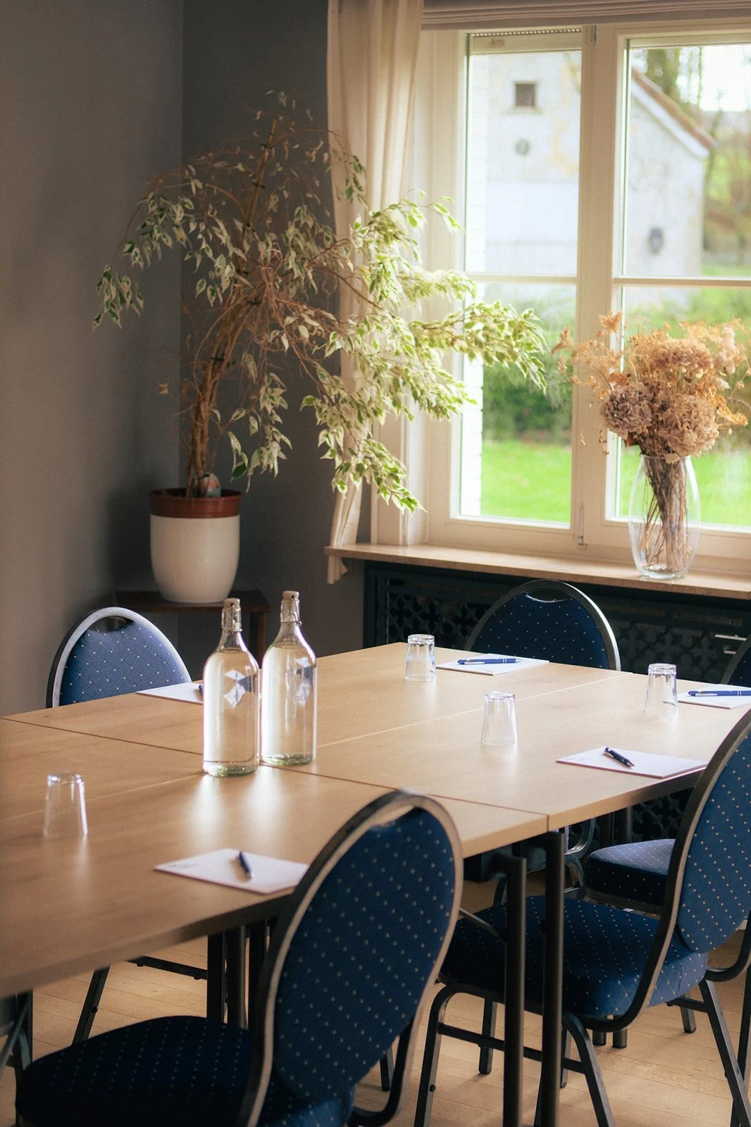 Salle de réunion avec une table en bois, six chaises avec assise bleue, deux bouteilles d'eau, six verres en plastique, six carnets avec stylos. Une grande fenêtre avec un rideau blanc laisse entrer la lumière naturelle. Sur la fenêtre, il y a un vas