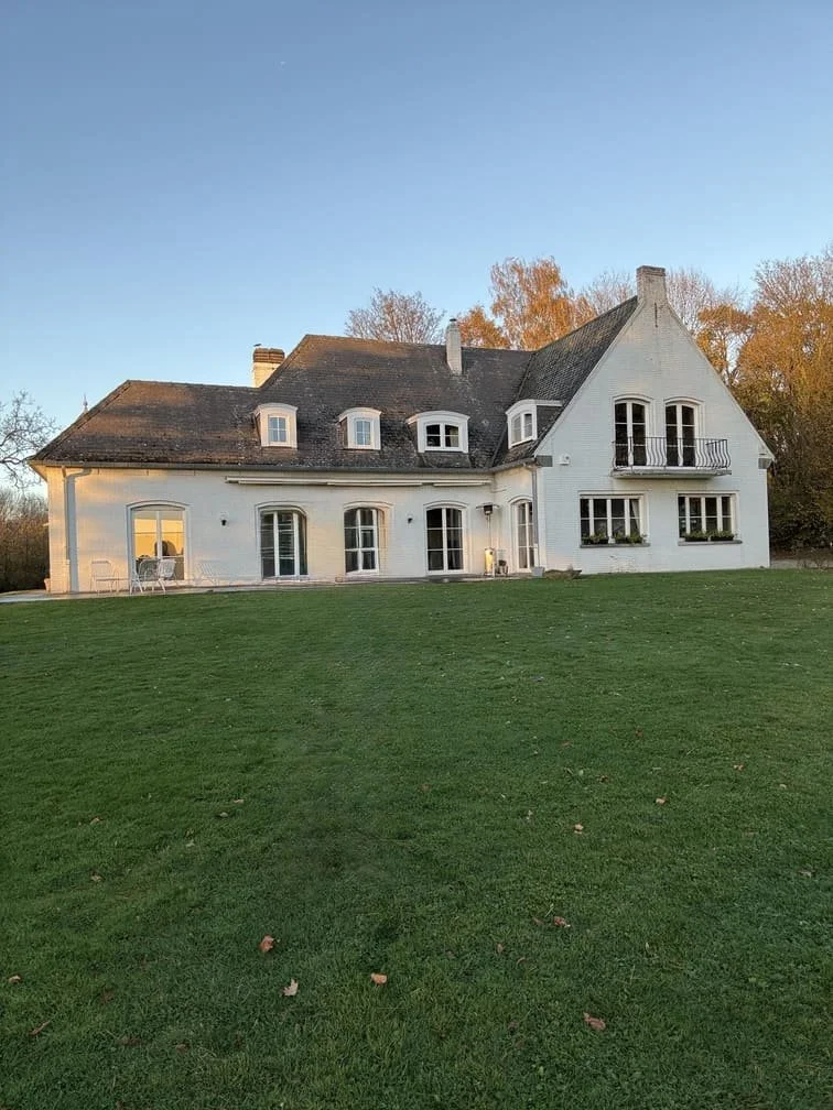 Une maison blanche à deux étages avec un toit en pente, plusieurs fenêtres, un balcon, et une grande pelouse verte devant elle sous un ciel clair.