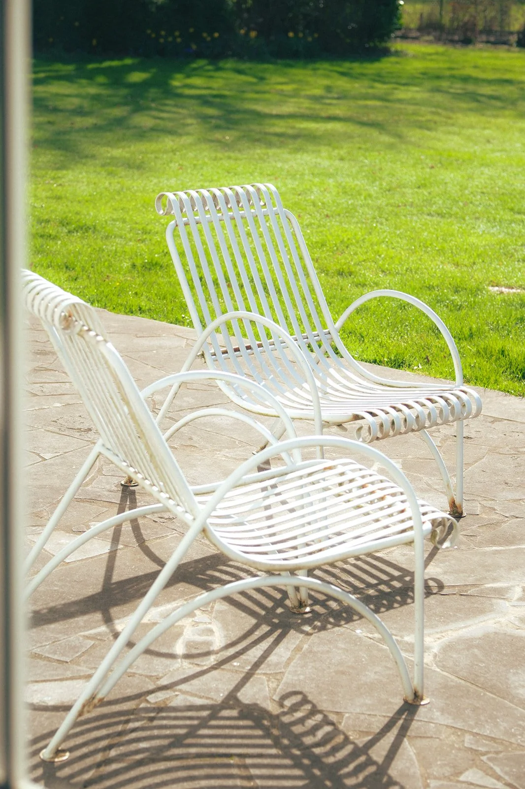 Deux chaises blanches en métal sur une terrasse en pierre avec un jardin vert en arrière-plan.