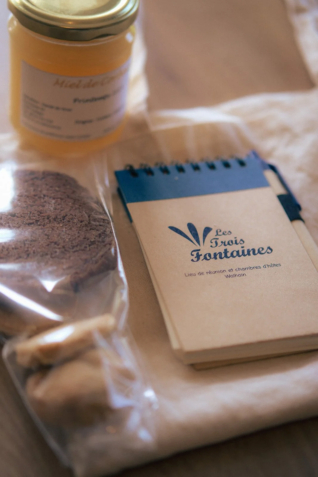 Exemple de goodie bug: Un carnet beige "Les Trois Fontaines" et une description indiquant un lieu de réunion et de chambres d'hôtes à Walhain, un sachet de biscuits locaux, miel, posés sur un sac en tissu beige.