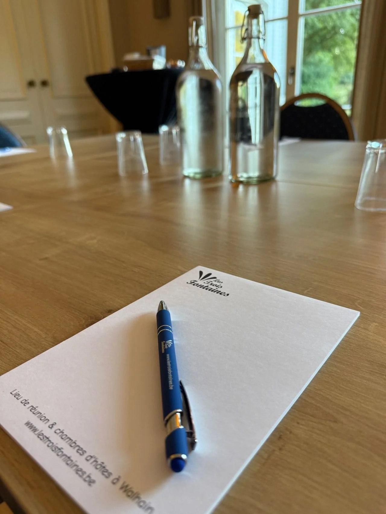 Une feuille de papier avec un stylo bleu dessus, sur une table en bois. En arrière-plan, deux bouteilles en verre et plusieurs verres. Fenêtre avec vue sur des arbres.
