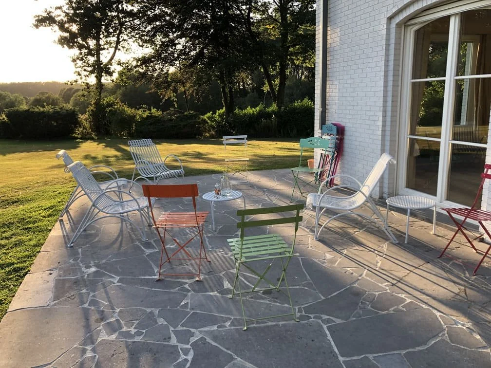 Terrasse avec plusieurs chaises colorées en métal, dehors en fin d'après-midi, avec un jardin verdoyant en arrière-plan et un bâtiment blanc à côté.