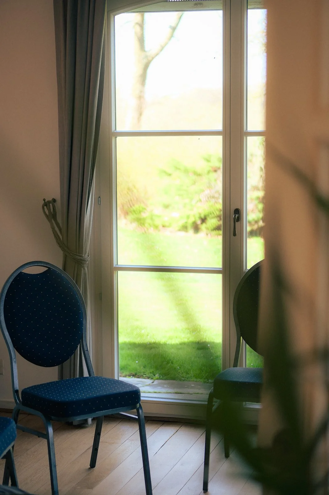 Une porte-fenêtre donnant sur un jardin vert, avec des chaises bleues autour, dans une pièce avec un sol en bois.