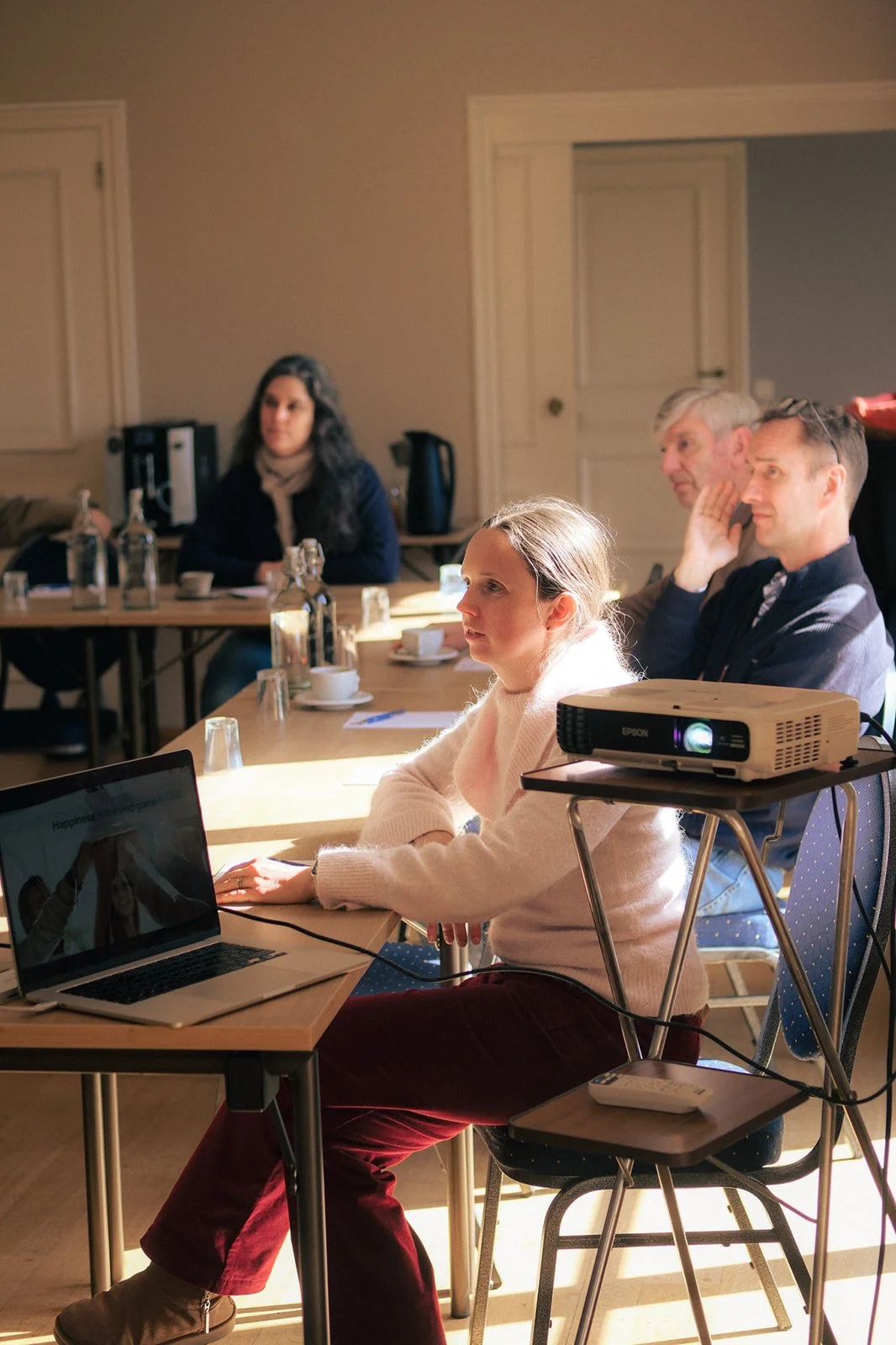 Groupe de personnes assises autour d'une table lors d'une réunion, projecteur, lumière naturelle dans une pièce. Les personnes ont l'air de bien se sentir dans la salle de réunion.