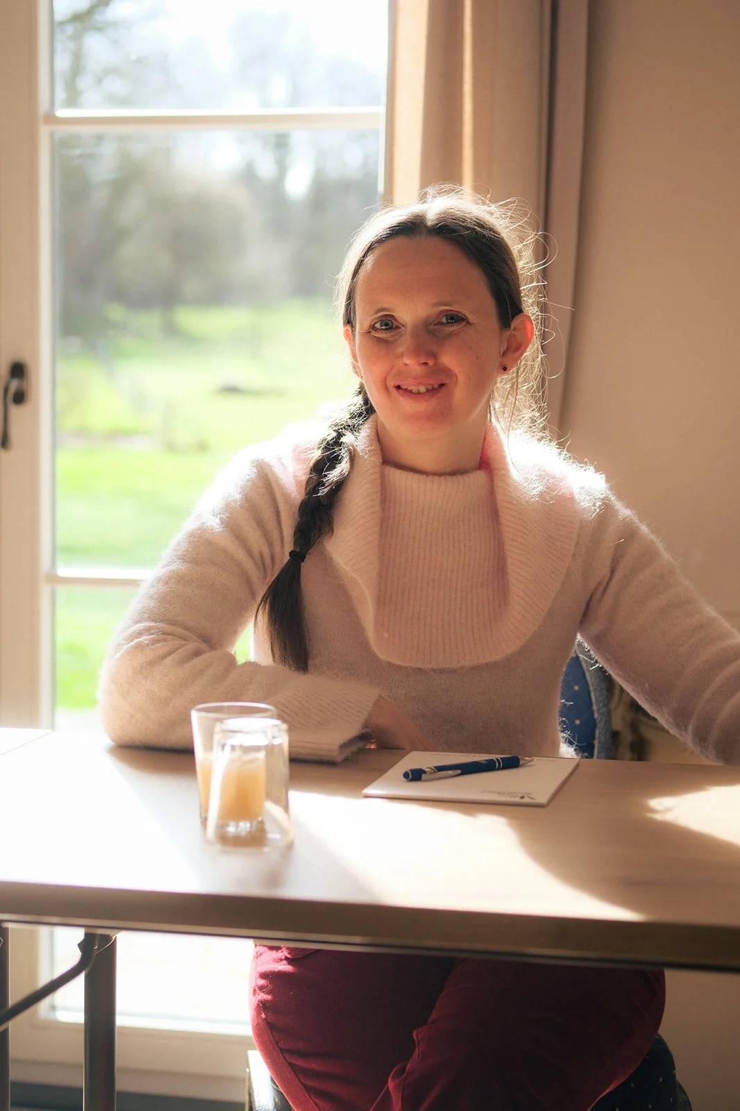 Une femme assise à une table près d'une fenêtre, souriante, avec un carnet, un stylo et un verre de boisson devant elle, éclairée par la lumière naturelle.