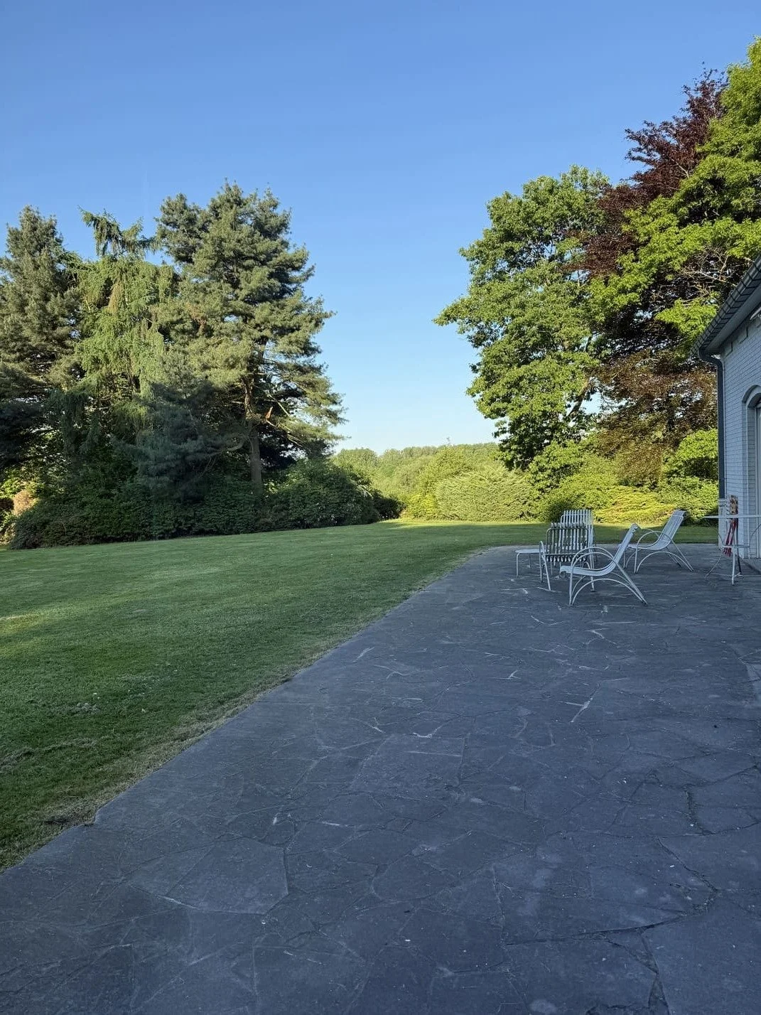 Une terrasse en pierre avec des chaises en métal, bordée d'une pelouse verte et d'arbres sous un ciel bleu clair.