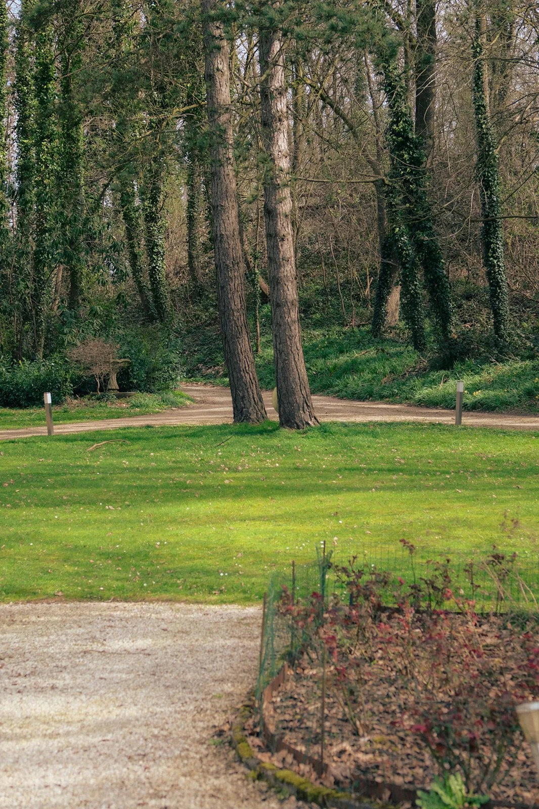 Le chemin en gravier amène à la villa des Trois Fontaines et bifurque dans une zone boisée avec des arbres verts et de la végétation dense. Espace de parking disponible.