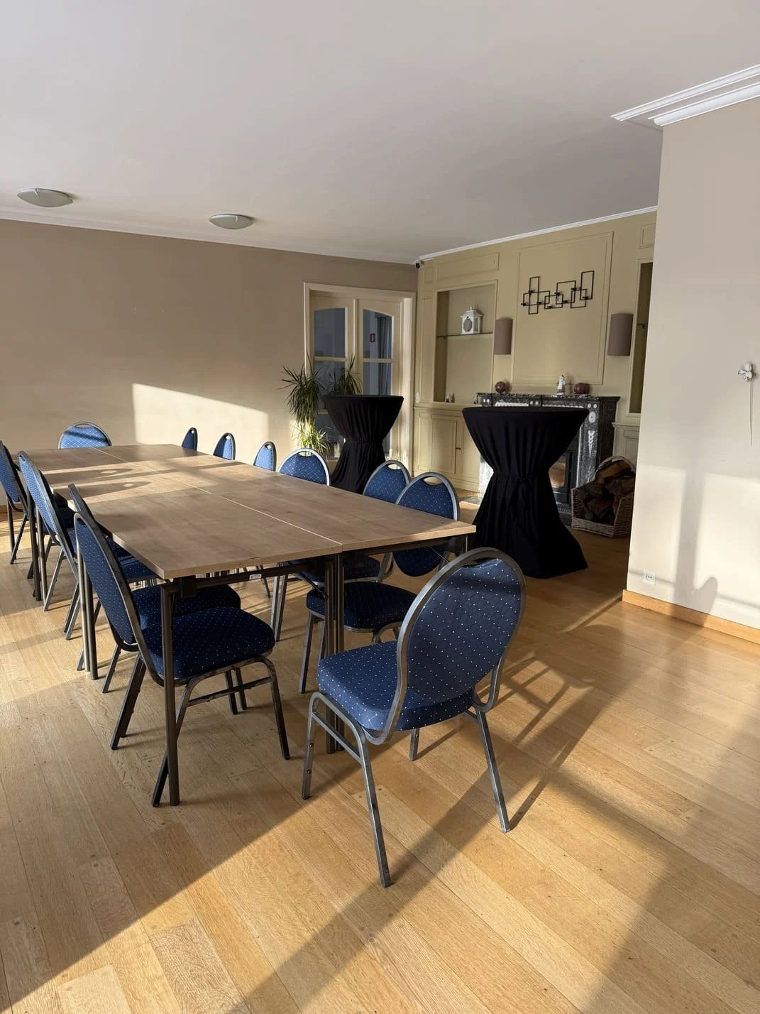 Salle de réunion avec une grande table en bois, chaises bleues avec arcades, et tables hautes avec housses noires, mur beige, cheminée avec décor, et plancher en bois.