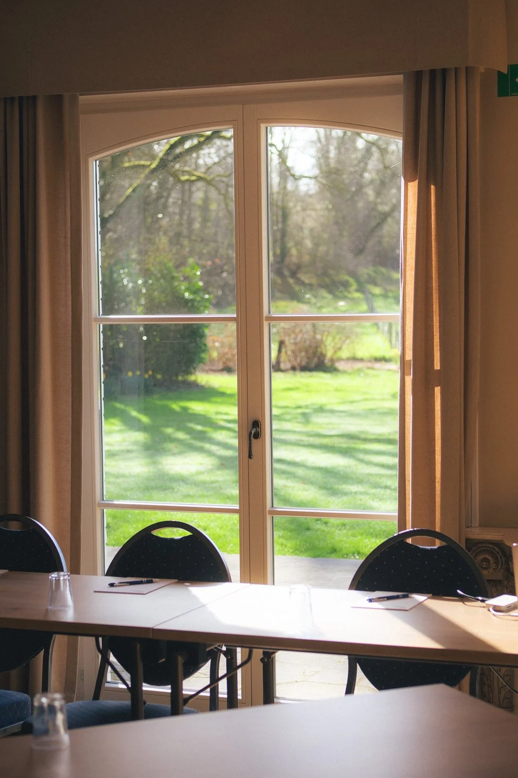 Une vue depuis l'intérieur d'une salle de réunion avec une grande fenêtre donnant sur un jardin ensoleillé, avec des chaises autour d'une table.