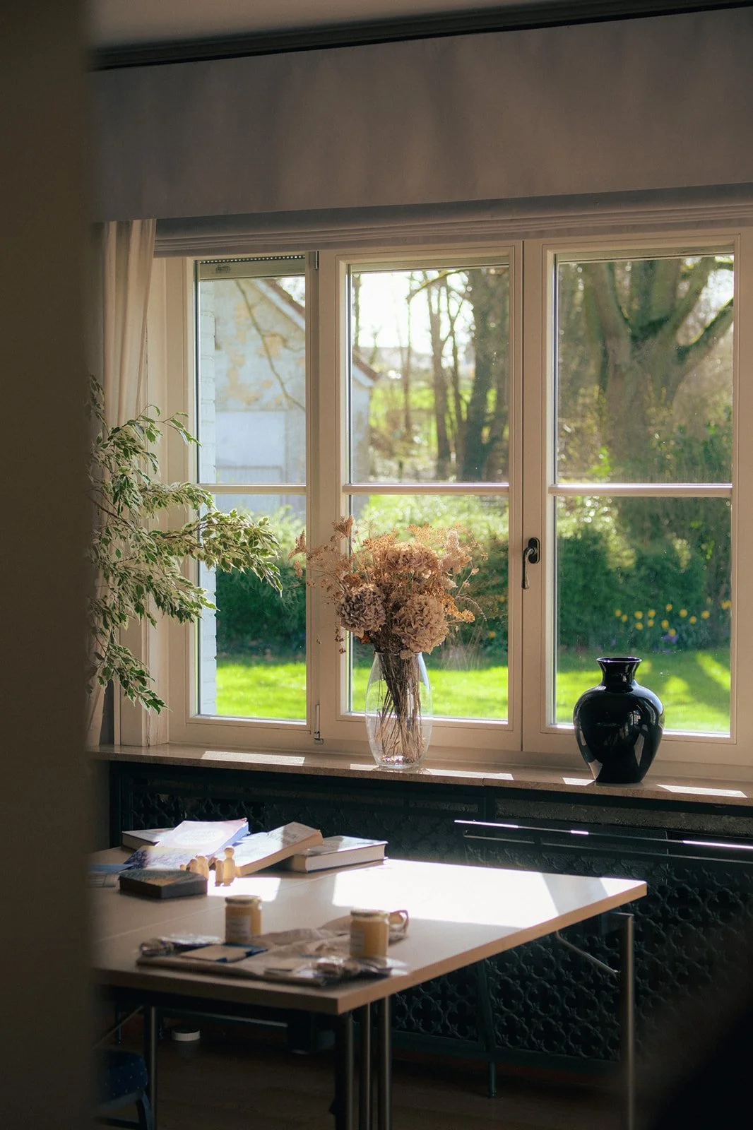 Intérieur d'un espace de réunion avec une fenêtre qui donne sur un jardin vert. Sur le rebord de la fenêtre, une vase en verre avec des fleurs séchées et une vase noire brillante. 