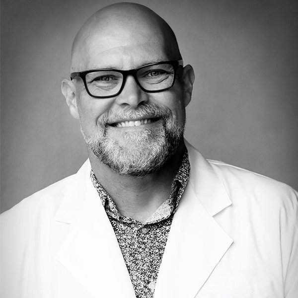 Black and white portrait of a smiling man with glasses, a beard, and a shaved head, wearing a white coat and a patterned shirt.