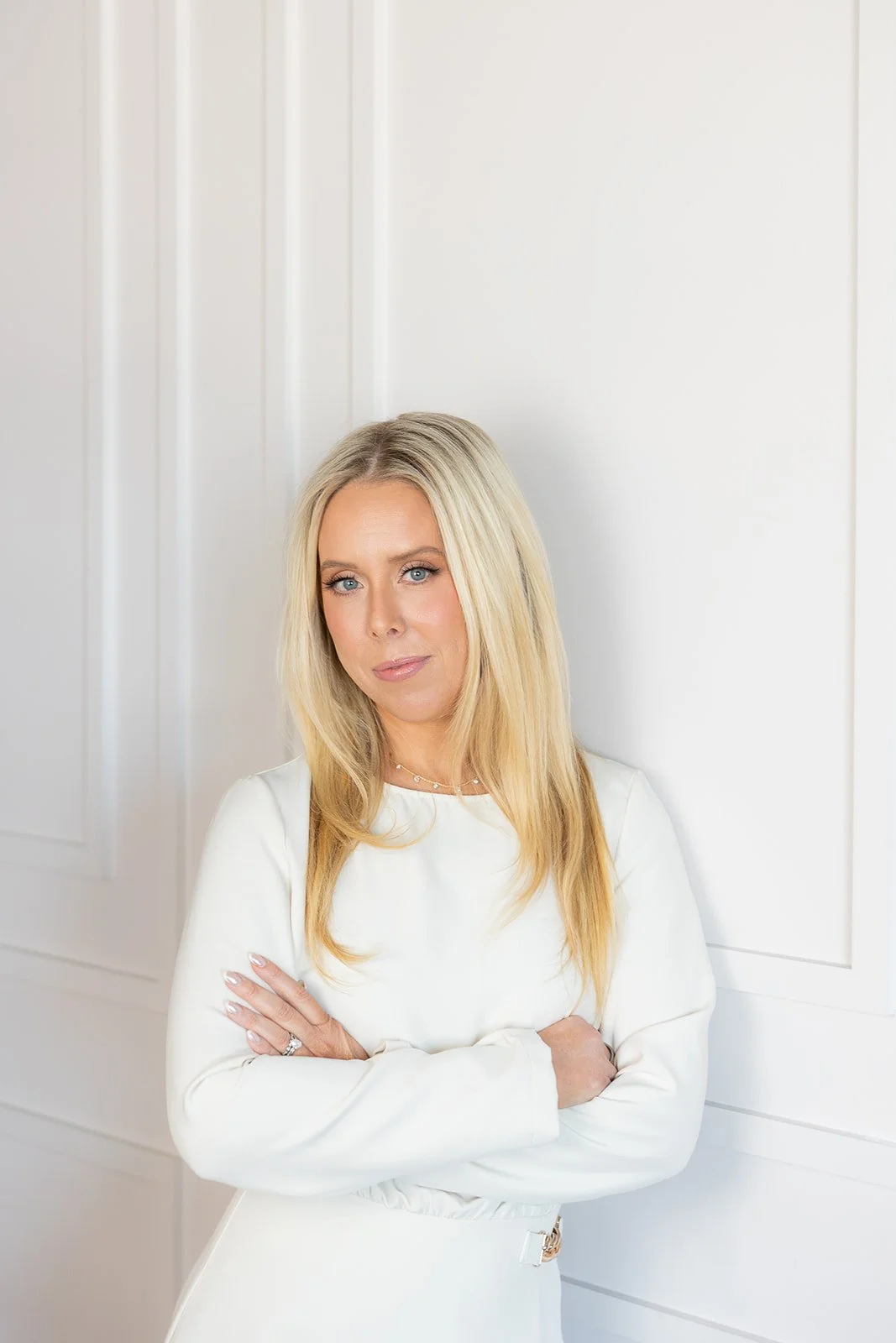 A woman with long blonde hair and blue eyes wearing a white long-sleeved shirt standing against a white wall with faint paneling, her arms crossed, looking at the camera.