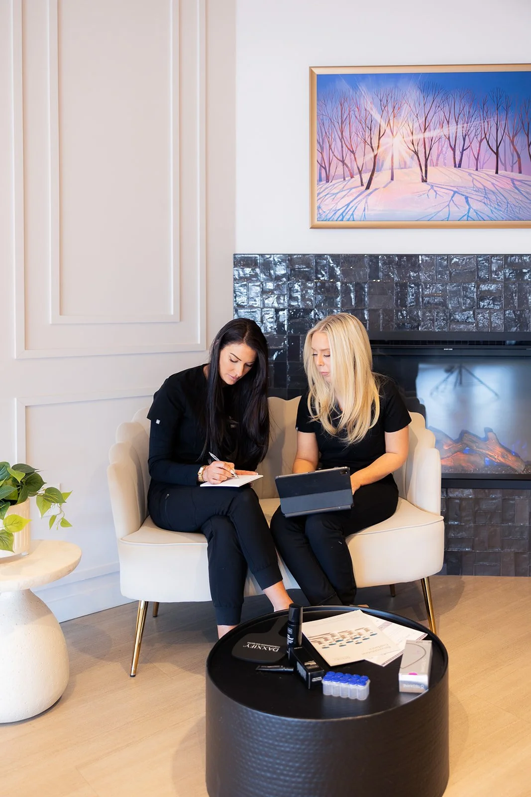 Two women sitting on a cream-colored sofa in a modern living room, looking at a tablet and writing notes. A black coffee table in front holds papers, a phone, and small containers. A black tiled fireplace is behind them, topped with a colorful painting of a winter scene with trees and sunlight.