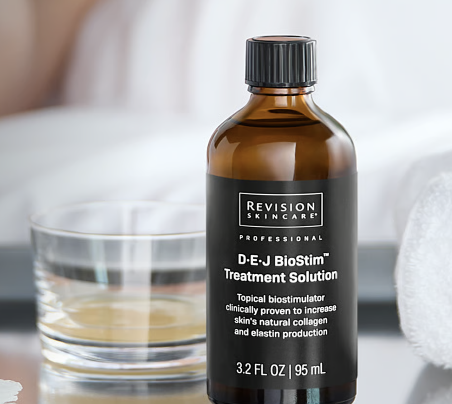 A brown bottle of Revision Skincare D.E.J BioStim Treatment Solution with a black label, next to a glass bowl filled with a clear liquid, on a grey surface with a soft white background.